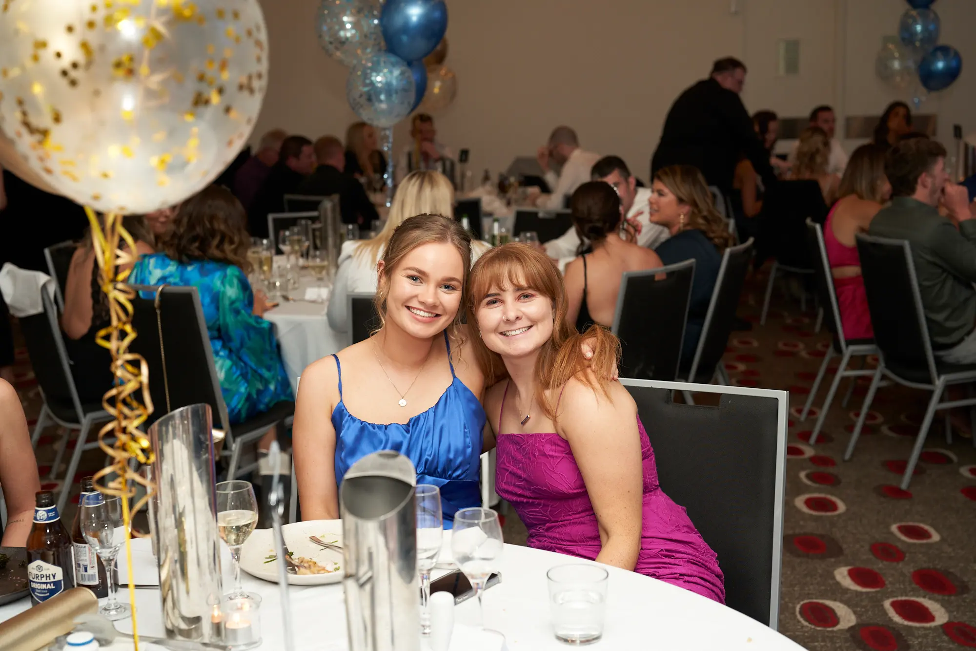 st kevins awards night pullman melbourne albert park two women smiling dinner table Two young women smile brightly for the camera while seated together at a dinner table during St. Kevin’s Awards Night at Pullman Melbourne Albert Park. The woman on the left wears a blue satin spaghetti-strap dress, while the woman on the right wears a magenta dress and has her arm around her friend’s shoulder. Behind them, the banquet hall is filled with elegantly dressed guests, white table settings, and festive clusters of blue and white balloons, capturing the joyful and celebratory mood of the evening.