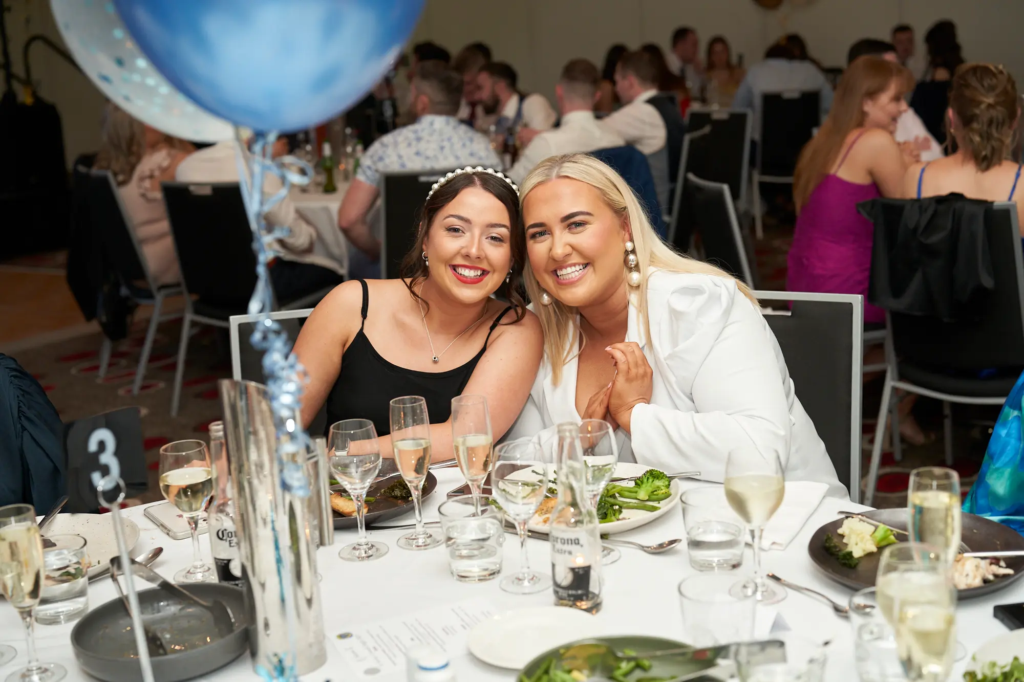 st kevins awards night pullman melbourne albert park two women smiling table 3 Two women smile warmly for the camera while seated together at a dinner table during St. Kevin’s Awards Night at Pullman Melbourne Albert Park. The woman on the left wears a black spaghetti-strap dress with a jeweled headband, and the woman on the right wears a white blazer. The table, marked with number “3,” is set with plates of food, glasses of wine, and bottles of Corona beer. In the background, guests dine in a crowded banquet hall decorated with blue, white, and silver balloons, capturing the joyful and festive atmosphere of the evening.