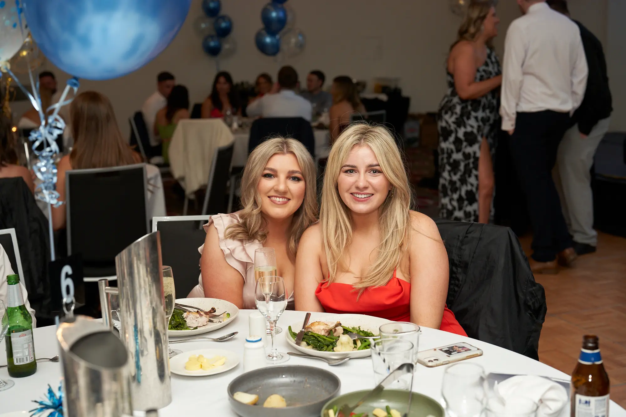 st kevins awards night pullman melbourne albert park two women smiling table number 6 Two women smile warmly for the camera while seated together at a dinner table during St. Kevin’s Awards Night at Pullman Melbourne Albert Park. The woman on the left wears a light pink ruffled dress, and the woman on the right wears a red strapless gown. Plates of food, drinks, and a table number “6” are visible in front of them. Behind them, elegantly dressed guests socialize in a lively banquet hall decorated with clusters of blue and gold balloons, reflecting the joyful and celebratory spirit of the evening.