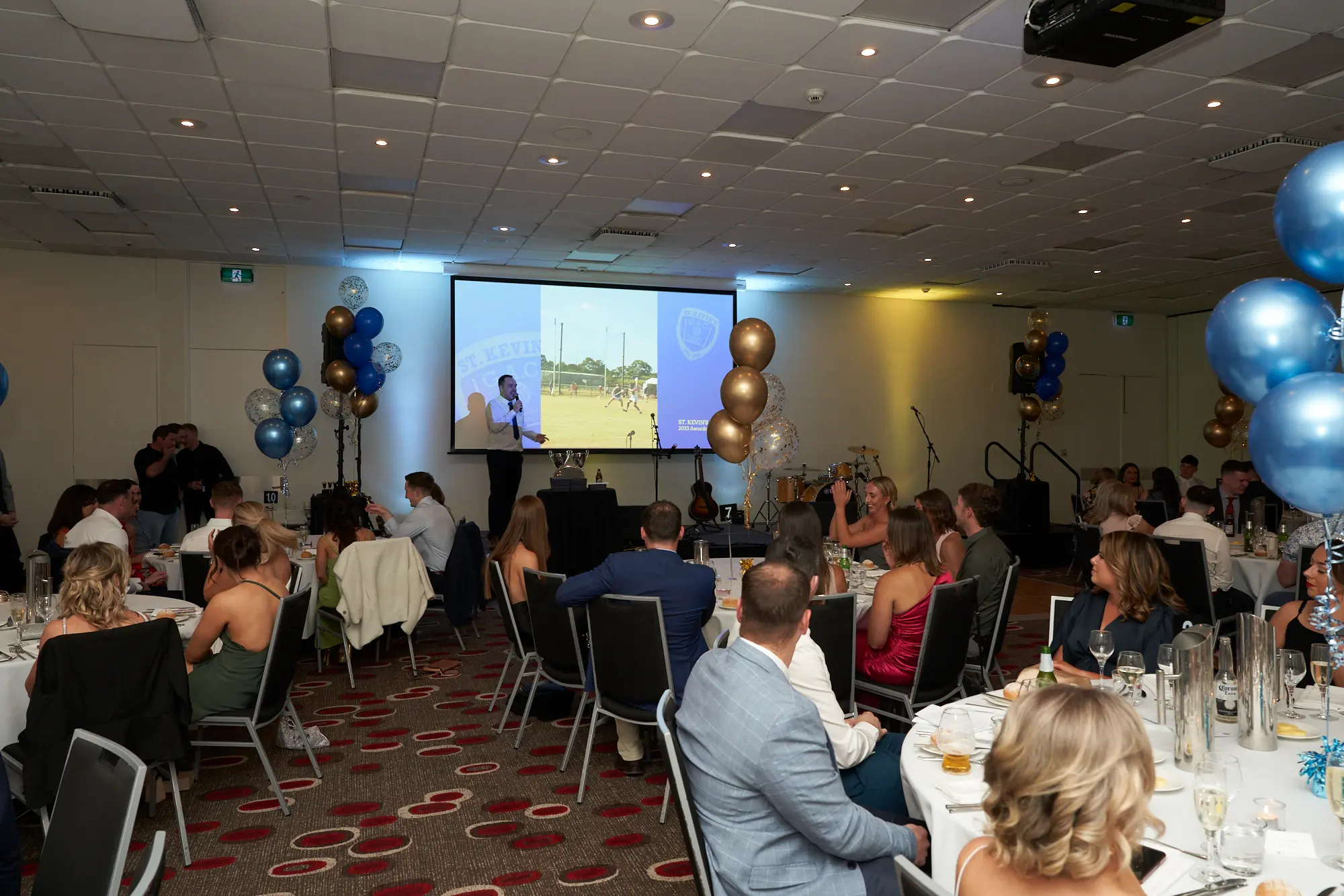 st kevins awards night pullman melbourne albert park wide shot stage speech A man stands at the center of a stage during St. Kevin's GAC Awards Night at Pullman Melbourne Albert Park, speaking into a microphone. Guests in formal attire are seated at round tables with white tablecloths, attentively listening. Blue, gold, and confetti-filled balloons decorate the room, while a large projector screen behind the speaker shows a sports scene. Musical instruments, including a drum set and speakers, flank the stage. The image captures the formal, festive, and social atmosphere of the awards night, highlighting both the engaged audience and the celebratory setting of this memorable event.