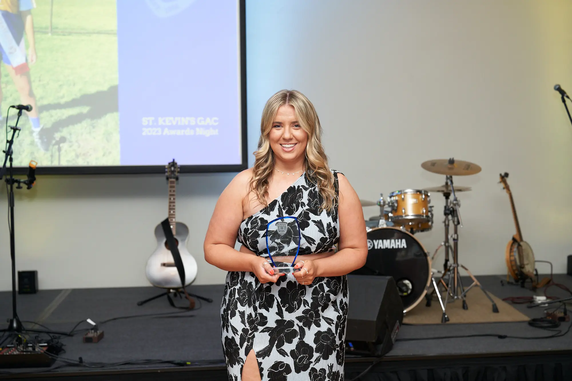 st kevins awards night pullman melbourne albert park woman floral award stage A woman poses proudly on stage at St. Kevin's GAC 2023 Awards Night at Pullman Melbourne Albert Park. She wears a black-and-white floral one-shoulder dress with a thigh-high slit and holds a clear glass award plaque. The stage features a guitar, drum set, additional string instrument, projection screen, and audio equipment, creating a formal and celebratory awards ceremony atmosphere.