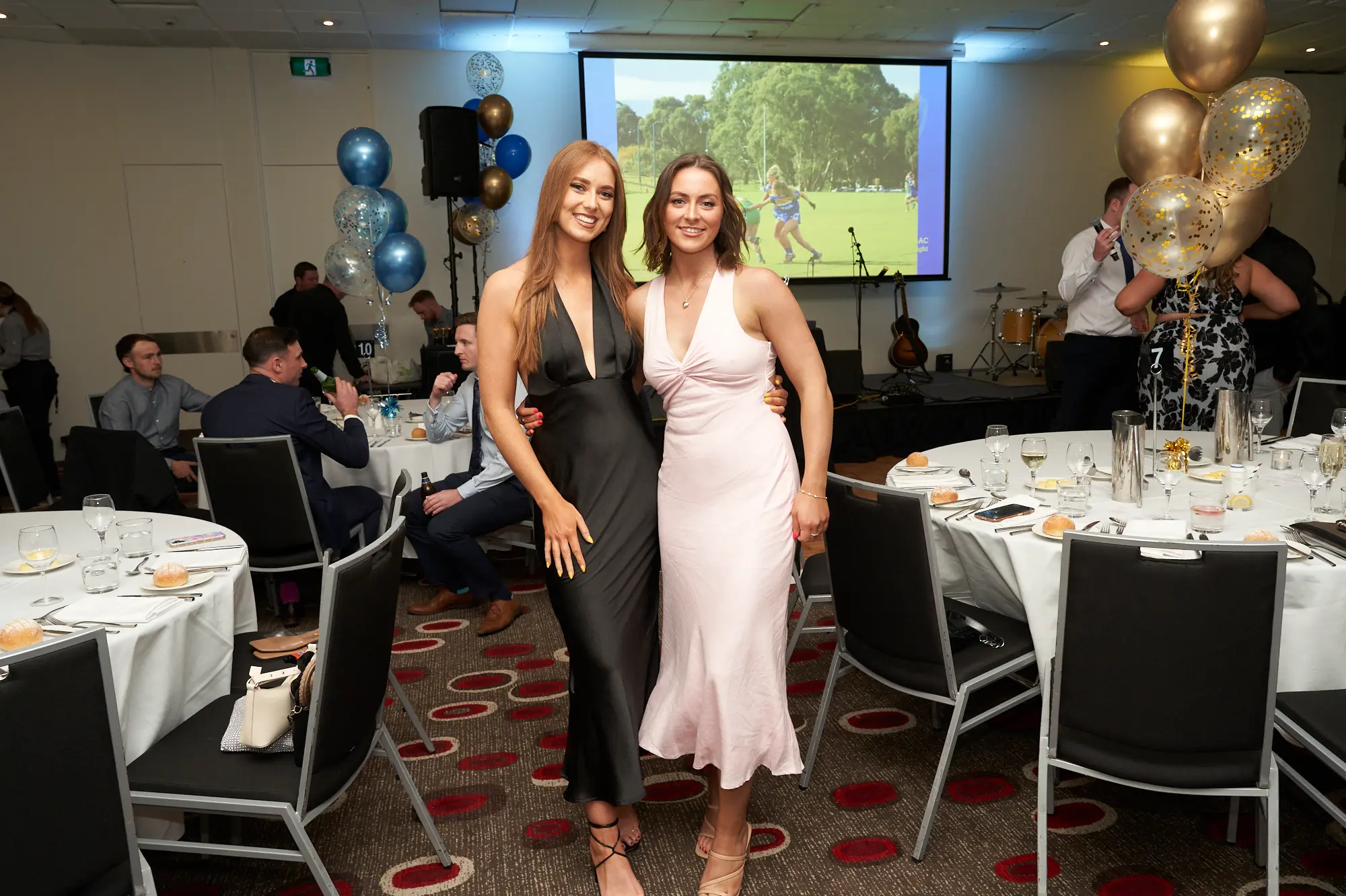 st kevins awards night pullman melbourne albert park women stage pose Two women stand side-by-side on a stage during the annual St. Kevin's Awards Night, smiling confidently at the camera. The blonde woman wears a black satin slip dress, while the dark-haired woman wears a light pink sleeveless dress. Behind them, a projection screen shows an outdoor sports scene, and musical instruments, including a guitar, cello/violin stand, and partial drum set, are arranged nearby. Gold, blue, and confetti balloons and a table with trophies decorate the stage. Guests seated at white-clothed tables in the background add to the formal, joyful, and celebratory atmosphere of the awards ceremony.