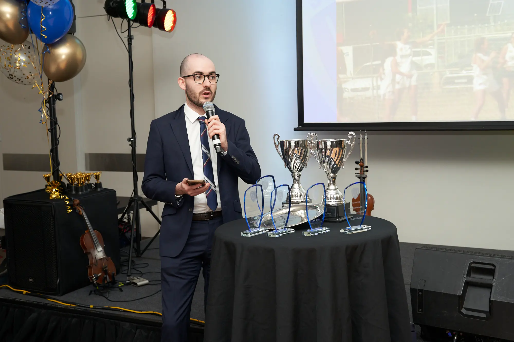 st kevins awards night speaker presenting on stage trophy table pullman melbourne albert park man in a dark suit, white shirt, and tie stands on stage at St. Kevin’s Awards Night, holding a microphone and a smartphone while delivering a speech. Next to him is a small round table draped in black, displaying trophies and awards. Behind him, a large screen projects an image of a sports team, and musical instruments including a cello and guitar are visible on the left side of the stage. Clusters of blue, gold, and white balloons add a festive touch. The atmosphere is formal and celebratory, highlighting a key award presentation moment at Pullman Melbourne Albert Park.