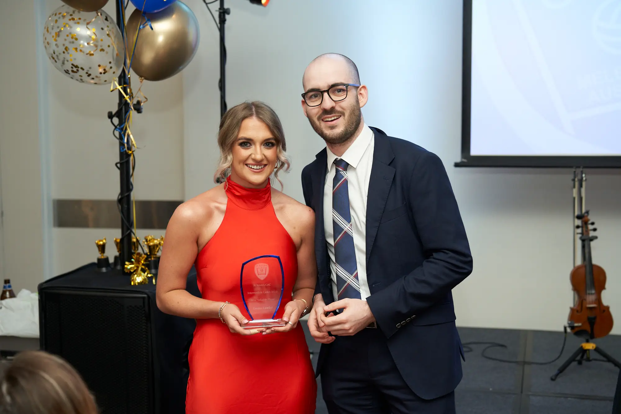 st kevins awards night winner holding glass plaque with presenter pullman melbourne albert park A woman in a vibrant red dress smiles while holding a glass award plaque beside a man in a dark suit and striped tie at St. Kevin’s Awards Night. Both are posing proudly for the camera, suggesting a recent award presentation. Behind them, a dark-covered table displays golden trophies, and festive balloons in gold, blue, and clear with confetti are tied to a stand. A large projector screen and a cello are also visible in the background. The atmosphere is formal, cheerful, and celebratory, reflecting the joy of an awards ceremony at Pullman Melbourne Albert Park.