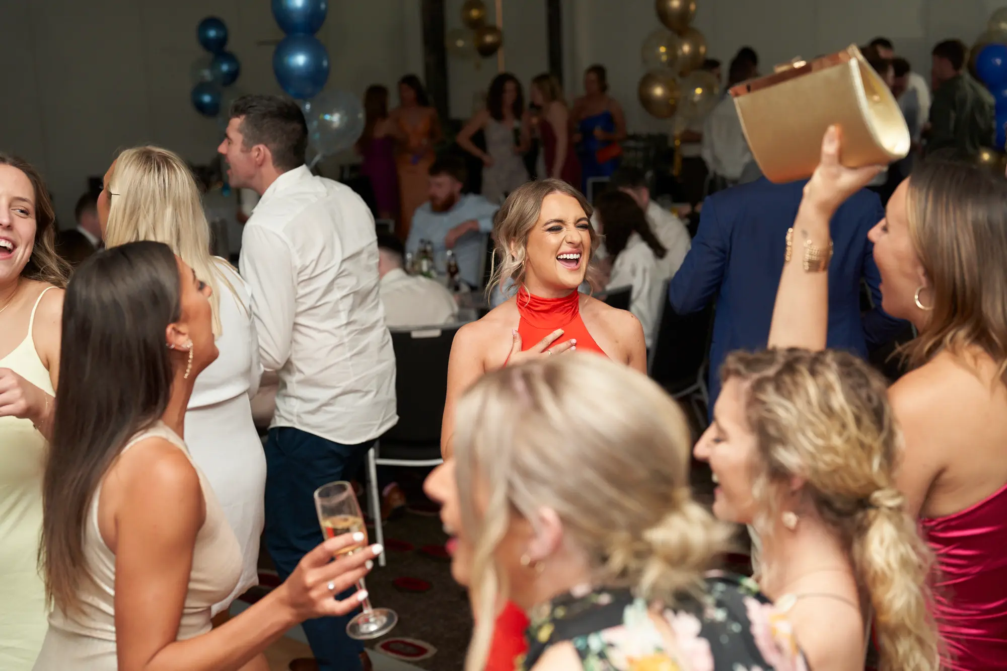 t kevins awards night pullman melbourne albert park dance floor laughter This wide shot captures a lively moment on the dance floor during the St. Kevin's Awards Night. A blonde woman in a vibrant red satin halter-neck dress laughs heartily, surrounded by friends in formal attire who are also smiling and enjoying themselves. Guests hold champagne flutes and a gold clutch, engaging in candid conversation and celebration. Festive gold, blue, and confetti balloons hover near the ceiling, while round tables with white tablecloths and black chairs fill the background. The scene radiates high energy, joy, and a vibrant post-awards party atmosphere.
