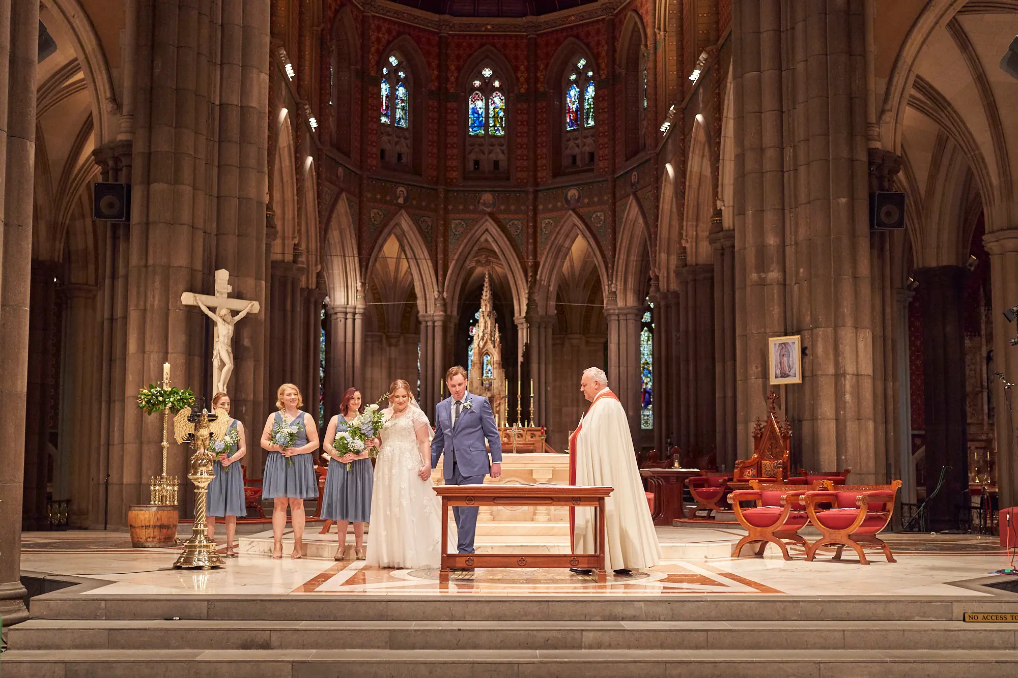 emily declan wedding altar bridesmaids st patricks cathedral melbourne The bride in a white lace dress and groom in a blue suit stand at a small wooden table on the altar of St Patrick’s Cathedral Melbourne, accompanied by four bridesmaids in dusty blue dresses holding bouquets, and a clergyman in red and white ceremonial vestments. They are posed on the raised platform in front of the altar. The Gothic Revival cathedral interior features soaring bluestone columns, high arches, ornate stained-glass windows, a large crucifix, red and white marble patterned floor, floral arrangements on wooden barrels, and traditional red chairs. The scene conveys solemnity, tradition, and bright reverence during the ceremony.