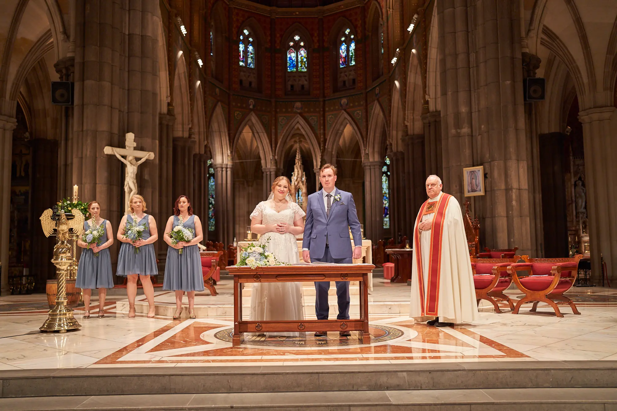 emily declan wedding altar group ceremony st patricks cathedral melbourne The bride in a white lace dress and groom in a blue suit stand at the altar of St Patrick’s Cathedral Melbourne, accompanied by three bridesmaids in dusty blue dresses holding white and blue bouquets, and a clergyman in red and white ceremonial vestments. They are positioned behind a wooden table on the raised platform. The Gothic Revival cathedral interior features soaring bluestone columns, high arches, ornate stained-glass windows, a large crucifix, red and white marble patterned floor, floral arrangements on wooden barrels, and traditional red chairs. The scene conveys solemnity, tradition, and bright reverence during the ceremony.