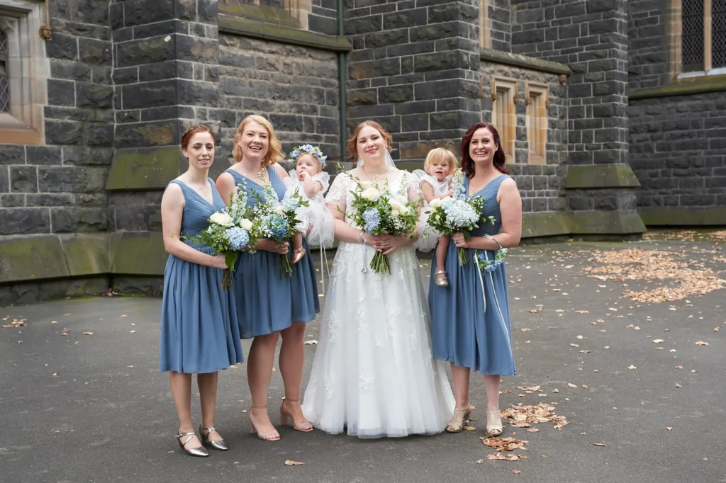 emily declan wedding bridal party children family st patricks cathedral A joyful bridal party and family posed in front of St Patrick’s Cathedral in Melbourne. The bride, in a white lace gown with cap sleeves and a V-neck, holds a white and blue bouquet and smiles warmly, while the groom, in a blue suit and brown shoes, has his arm around her. Four bridesmaids in matching dusty or steel blue knee-length V-neck dresses stand alongside them, holding similar bouquets, with one playfully looking at the baby flower girl. The baby flower girl in a white dress with a floral headband is held on the left, and a young page boy in a suit stands next to the groom. They are set against the dark rock-faced bluestone cathedral exterior with ornate windows and sandstone trim, creating a celebratory, elegant, and grand portrait.