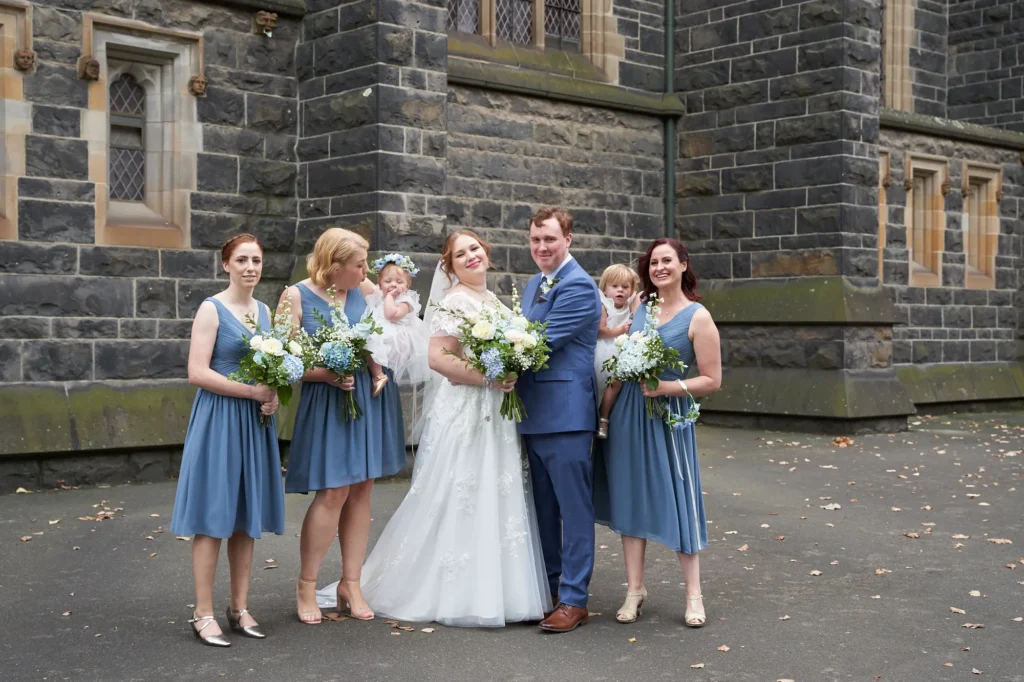 emily declan wedding bridal party family portrait st patricks cathedral A joyful bridal party and family posed in front of St Patrick’s Cathedral in Melbourne. The bride, wearing a white lace gown with cap sleeves and a V-neck, holds a white and blue bouquet and smiles, while the groom, in a blue suit and brown shoes, has his arm around her. Four bridesmaids in matching dusty blue or steel blue knee-length V-neck dresses stand alongside them, holding similar bouquets. A baby flower girl in a white dress with a floral headband is held on the left, and a young page boy in a suit stands next to the groom. The group is set against the dark rock-faced bluestone Gothic Revival cathedral exterior with ornate windows and sandstone trim, creating a celebratory and elegant portrait.