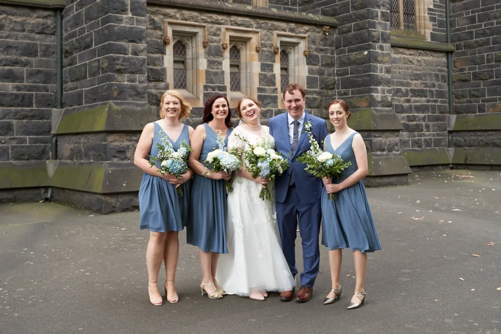 emily declan wedding bridal party joyful portrait st patricks cathedral A joyful bridal party posed in front of St Patrick’s Cathedral in Melbourne. The bride, wearing a white lace gown with cap sleeves and a V-neck, holds a vibrant white and blue bouquet and laughs joyfully with the groom, who wears a blue suit and patterned tie. Four bridesmaids in matching dusty blue or steel blue knee-length V-neck dresses stand alongside them, holding similar bouquets and smiling at the camera. They are posed against the dark rock-faced bluestone Gothic Revival cathedral exterior with tall narrow windows and sandstone trim, creating a celebratory, elegant, and timeless portrait.