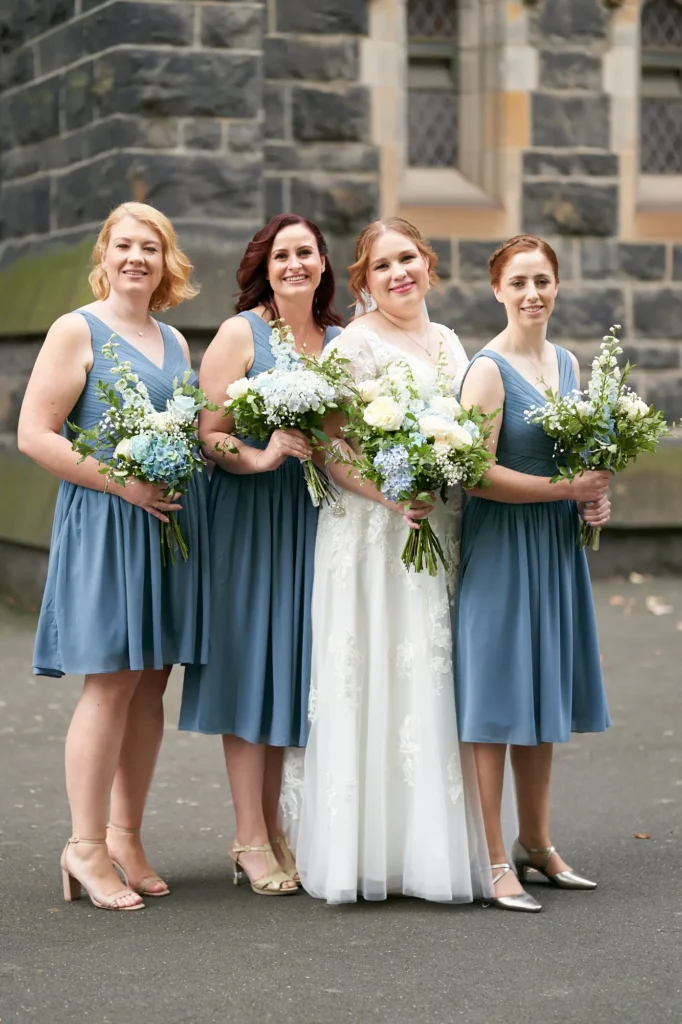 emily declan wedding bridal party timeless portrait st patricks cathedral melbourne The bride and her three bridesmaids are posed in front of St Patrick’s Cathedral Melbourne for a joyful bridal party portrait. The bride, wearing a white wedding gown with lace cap sleeves and a V-neck, holds a white and blue bouquet and smiles warmly at the camera. The bridesmaids, dressed in matching dusty blue knee-length dresses with V-neck bodices, flank her holding similar bouquets. They stand against the dark, rock-faced bluestone exterior of the Gothic Revival cathedral, with ornate stone arches and stained-glass windows visible in the background, creating a celebratory, elegant, and timeless group portrait.