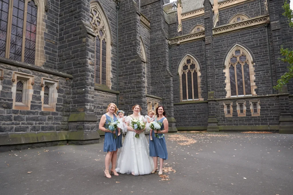 emily declan wedding bridal party toddler st patricks cathedral A joyful bridal party posed in front of St Patrick’s Cathedral in Melbourne. The bride, in a white lace gown with cap sleeves and a V-neck, holds a white and blue bouquet and smiles warmly at the camera. Three bridesmaids in matching dusty or steel blue knee-length V-neck dresses stand alongside her, holding similar bouquets. A young toddler in a white dress with a floral headband stands next to the bridesmaid on the far right. The group is set against the dark rock-faced bluestone exterior of the cathedral with tall ornate windows and sandstone trim, creating a celebratory, elegant, and grand portrait.