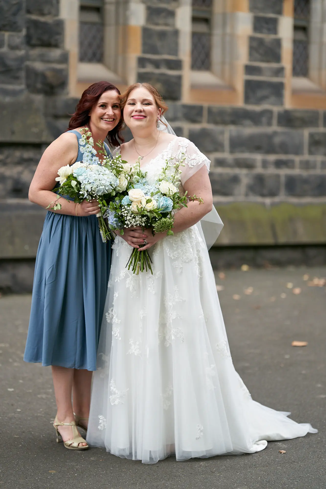 emily declan wedding bride and bridesmaid happy portrait st patricks cathedral A joyful moment between the bride and her bridesmaid in front of St Patrick’s Cathedral in Melbourne. The bride, on the right, wears a white lace wedding gown with cap sleeves and a V-neck, holding a vibrant white and blue bouquet, smiling warmly. The bridesmaid, on the left, wears a dusty blue V-neck dress, holds a similar bouquet, and has her arm around the bride. They are posed against the dark, rock-faced bluestone Gothic Revival exterior of the cathedral, with an ornate window and sandstone trim in the background, creating an elegant, celebratory, and timeless image.