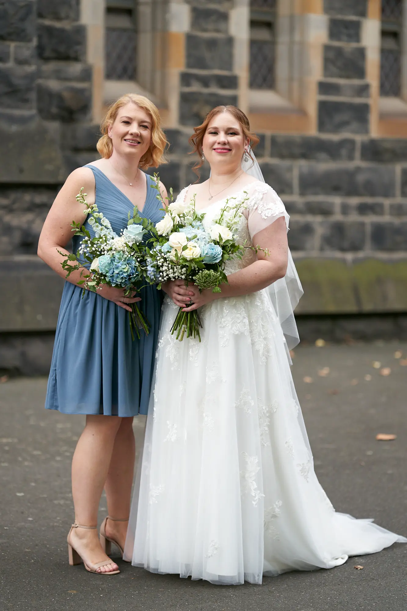 emily declan wedding bride and bridesmaid joyful portrait st patricks cathedral The bride and a bridesmaid are posed in front of St Patrick’s Cathedral in Melbourne for a joyful portrait. The bride, wearing a white wedding gown with lace cap sleeves and a V-neck, stands on the left holding a vibrant white and blue bouquet, smiling warmly at the camera. The bridesmaid, in a matching dusty blue knee-length V-neck dress with metallic silver shoes, stands to the right holding a similar bouquet. They are set against the dark, rock-faced bluestone exterior of the Gothic Revival cathedral, with large windows and sandstone trim in the background, creating an elegant and celebratory image.