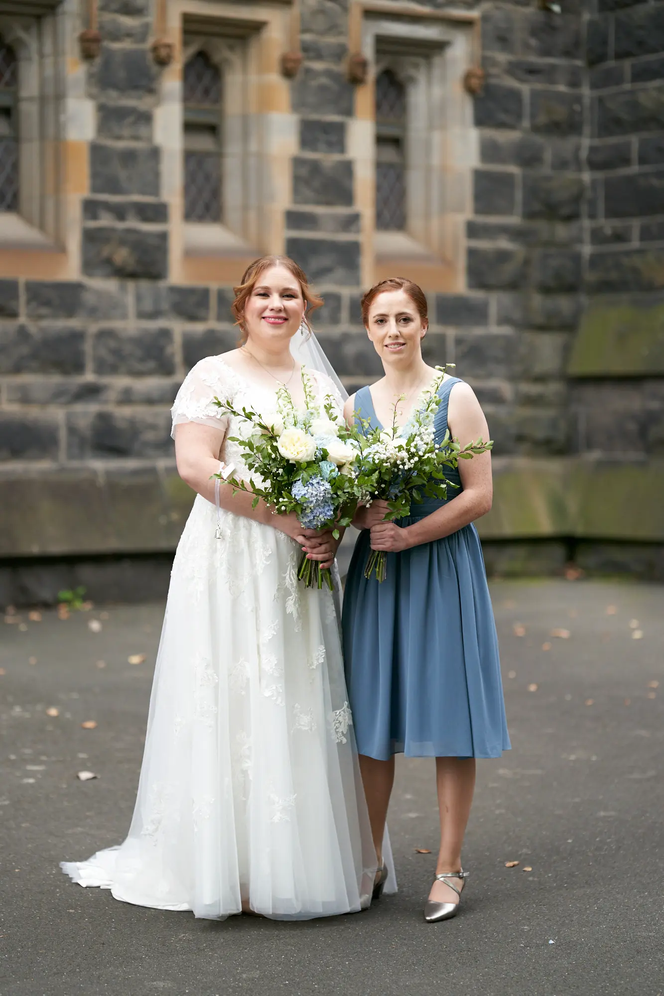 emily declan wedding bride and bridesmaid portrait st patricks cathedral melbourne The bride and a bridesmaid are posed in front of St Patrick’s Cathedral Melbourne for a joyful portrait. The bride, wearing a white wedding gown with lace cap sleeves and a V-neck, stands on the left holding a vibrant white and blue bouquet, smiling warmly at the camera. The bridesmaid, in a matching dusty blue knee-length V-neck dress and metallic silver shoes, stands to the right holding a similar bouquet. They are set against the dark, rock-faced bluestone exterior of the Gothic Revival cathedral, with ornate stone arches and large windows in the background, creating an elegant and celebratory image.