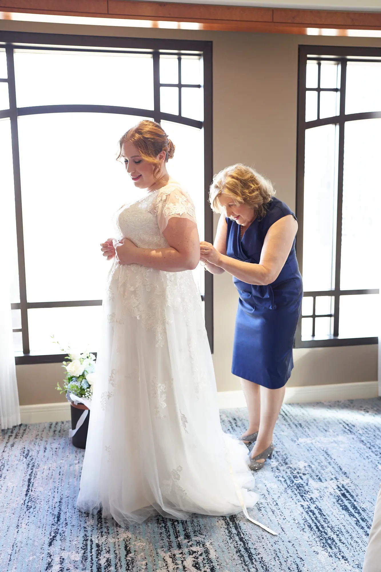 emily declan wedding bride assisted into gown park hyatt melbourne The bride stands in profile wearing her lace, cap-sleeved wedding dress at the Park Hyatt Melbourne, while a close family member in a navy blue dress fastens the back of the gown. Natural light floods the luxurious hotel suite through large windows, illuminating the textured blue and white carpet and a subtle floral arrangement. The bride’s calm, anticipatory expression and the helper’s focused care capture an intimate, heartfelt moment, reflecting the warmth, love, and quiet elegance of Emily and Declan’s pre-ceremony preparations.