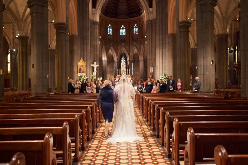 emily declan wedding bride entrance st patricks cathedral Emily, the bride, walks down the central aisle of St Patrick’s Cathedral Melbourne, accompanied by a loved one, in a long white gown with train and veil. Surrounded by towering Gothic Revival stone pillars, soaring arches, and stained-glass windows, the cathedral’s grandeur frames this solemn wedding moment. Guests seated in wooden pews watch as the couple approaches the brightly lit altar, where sunlight and interior lighting highlight intricate architectural details. The image captures timeless elegance, reverence, and anticipation, reflecting the profound significance of the ceremony within Australia’s largest church
