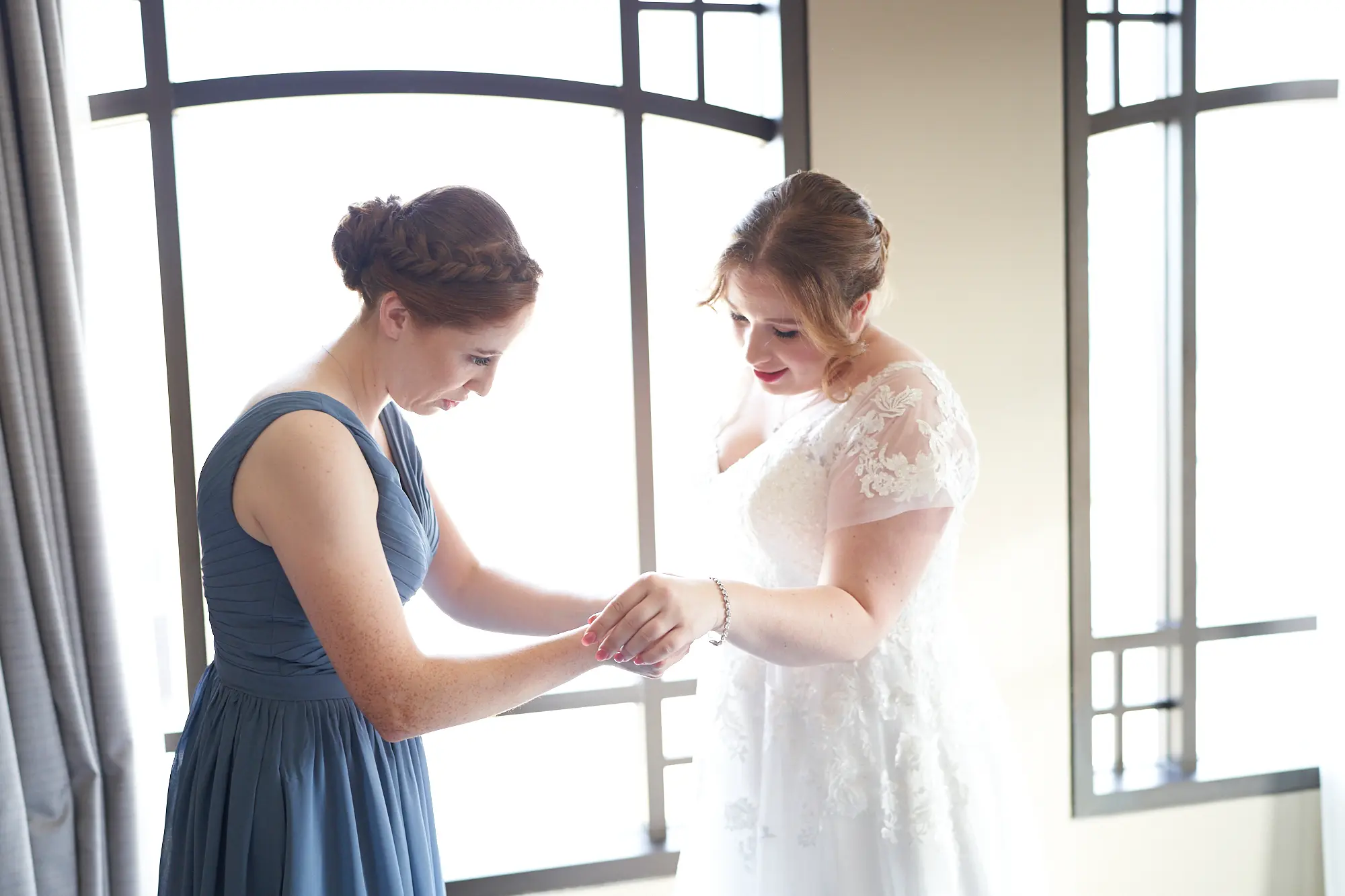 emily declan wedding bride getting ready bracelet park hyatt melbourne The bride in her white lace cap-sleeved wedding dress has a bracelet fastened by a close family member or friend in a slate-blue V-neck dress at the Park Hyatt Melbourne. Natural light floods the modern hotel suite through large windows, creating a soft, warm glow that highlights the delicate lace and intimate gesture. This tender, personal moment captures quiet love, support, and anticipation, reflecting the serene and heartfelt atmosphere of Emily and Declan’s pre-ceremony wedding preparations.