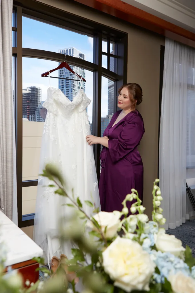 emily declan wedding bride getting ready park hyatt melbourne ride getting ready at the Park Hyatt Melbourne, standing near a large window in a flowing purple robe, gently adjusting her strapless white wedding dress. The luxurious hotel suite features neutral tones, plush carpet, and an elegant floral arrangement, while the window frames a striking view of Melbourne’s city skyline. Soft natural light illuminates the serene, anticipatory moment, capturing the calm and sophistication of the pre-wedding preparation. This image highlights the quiet elegance and reflective pause before the wedding ceremony, blending intimate indoor details with the dynamic urban backdrop.