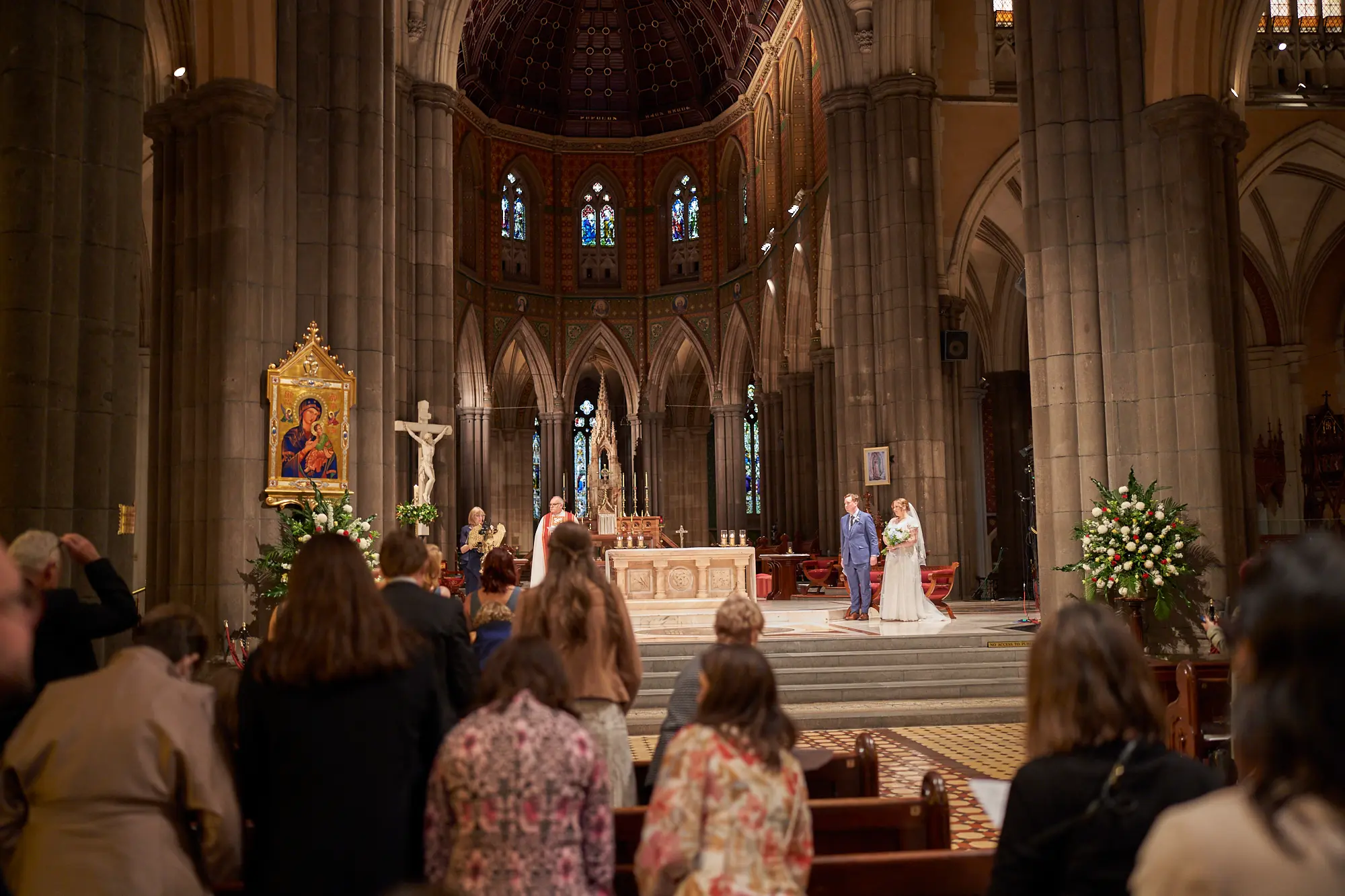 emily declan wedding bride groom altar ceremony st patricks cathedral melbourne The bride and groom stand at the altar of St Patrick’s Cathedral Melbourne during their wedding ceremony, facing a clergyman in white and red vestments presiding over the service. The bride wears a white lace gown and holds her bouquet, while the groom is in a blue suit. Guests are seated in the pews observing the ceremony. Soaring bluestone columns, high Gothic arches, ornate stained-glass windows, a mosaic-domed ceiling, and a white marble altar with a prominent crucifix frame the scene. This bright, solemn, and reverent moment captures the grandeur and timeless elegance of Emily and Declan’s wedding ceremony.