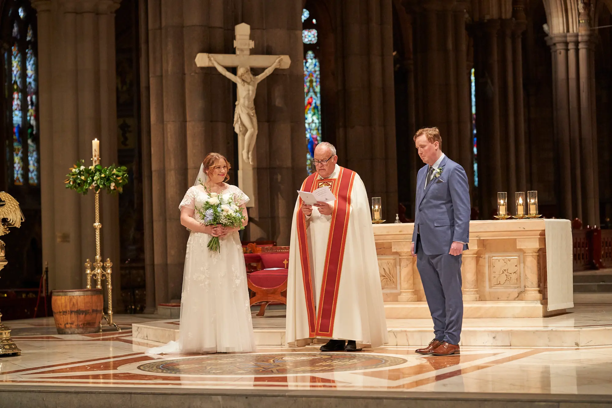 emily declan wedding bride groom ceremony altar st patricks cathedral melbourne The bride and groom stand at the altar of St Patrick’s Cathedral Melbourne during their wedding ceremony, facing a clergyman in white and red ceremonial vestments reading from a red book. The bride wears a white lace gown and holds her bouquet, while the groom is in a blue suit. They stand on a raised platform with a red and white marble patterned floor, beneath soaring bluestone columns and Gothic arches. Ornate stained-glass windows, a large crucifix, and floral arrangements frame the scene, capturing solemnity, grandeur, and bright reverence during Emily and Declan’s sacred wedding vows.