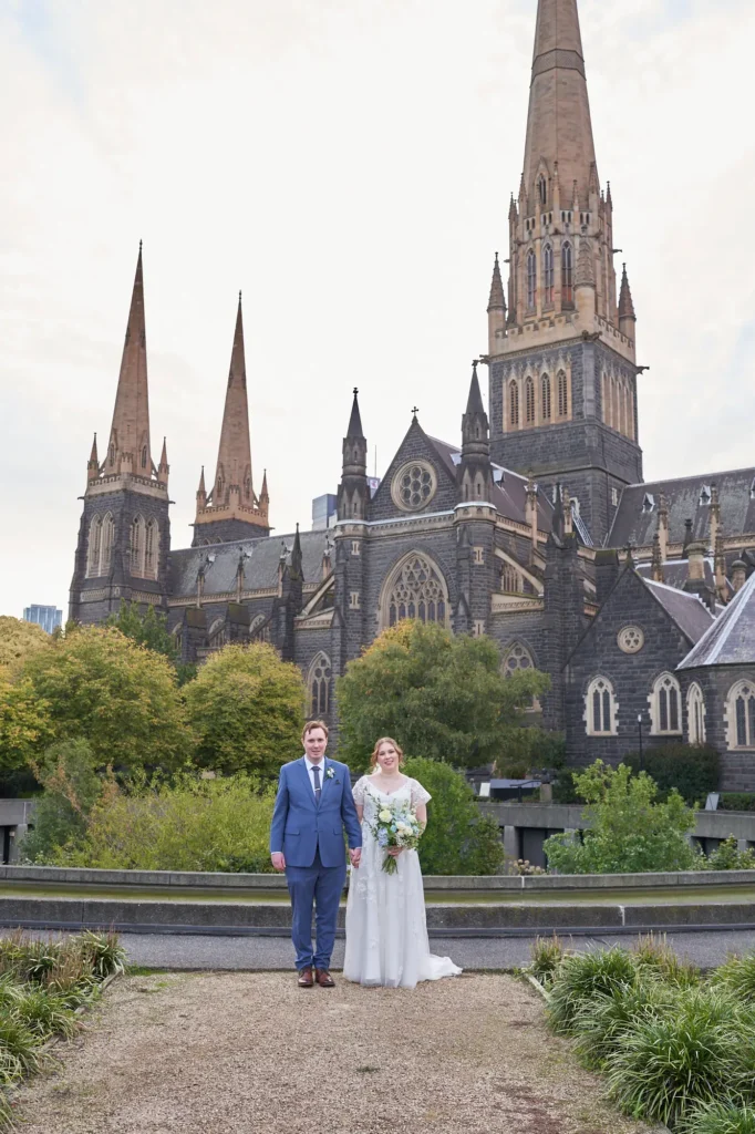 emily declan wedding bride groom courtyard st patricks cathedral A bride and groom pose for a formal portrait in front of St Patrick’s Cathedral in Melbourne. The groom wears a light blue suit and brown shoes, and the bride wears a white V-neck A-line lace wedding dress with cap sleeves, holding a white and blue bouquet. They stand hand-in-hand on a dark paved courtyard with low green foliage at their feet. The cathedral’s dark rock-faced bluestone exterior, intricate stonework, ornate windows, and soaring Gothic spires form a grand, historic backdrop under a soft overcast sky, creating a serene and timeless atmosphere.