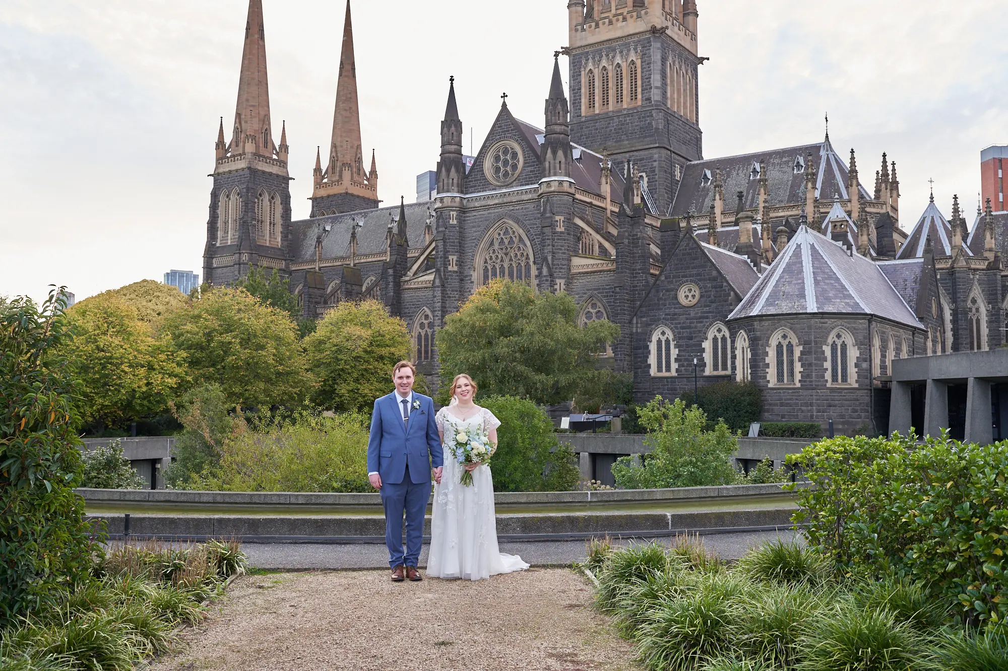 emily declan wedding bride groom formal courtyardst patricks cathedral A bride and groom pose for a formal portrait in front of St Patrick’s Cathedral in Melbourne. The groom wears a light blue suit and brown shoes, and the bride wears a white V-neck A-line lace wedding dress with cap sleeves, holding a white and blue bouquet. They stand hand-in-hand on a dark paved courtyard with low green foliage. The cathedral’s dark rock-faced bluestone exterior, intricate stonework, ornate windows, and soaring Gothic spires form a grand, historic backdrop under a soft overcast sky, creating a serene and timeless atmosphere.