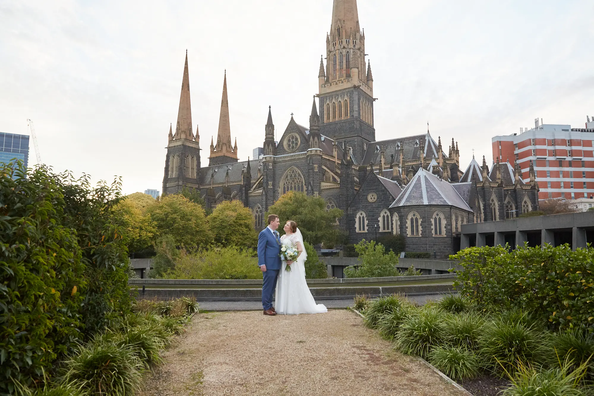 emily declan wedding bride groom garden courtyard st patricks cathedral A bride and groom share an intimate moment, holding hands and looking at each other in front of St Patrick’s Cathedral in Melbourne. The groom wears a light blue suit and brown shoes, and the bride wears a white V-neck A-line lace wedding dress with cap sleeves, holding a white and blue bouquet. They stand on a paved walkway in a garden area with low green foliage, framed by the cathedral’s dark rock-faced bluestone exterior, soaring Gothic spires, and overcast sky, highlighting the grandeur of the historic Gothic Revival architecture.
