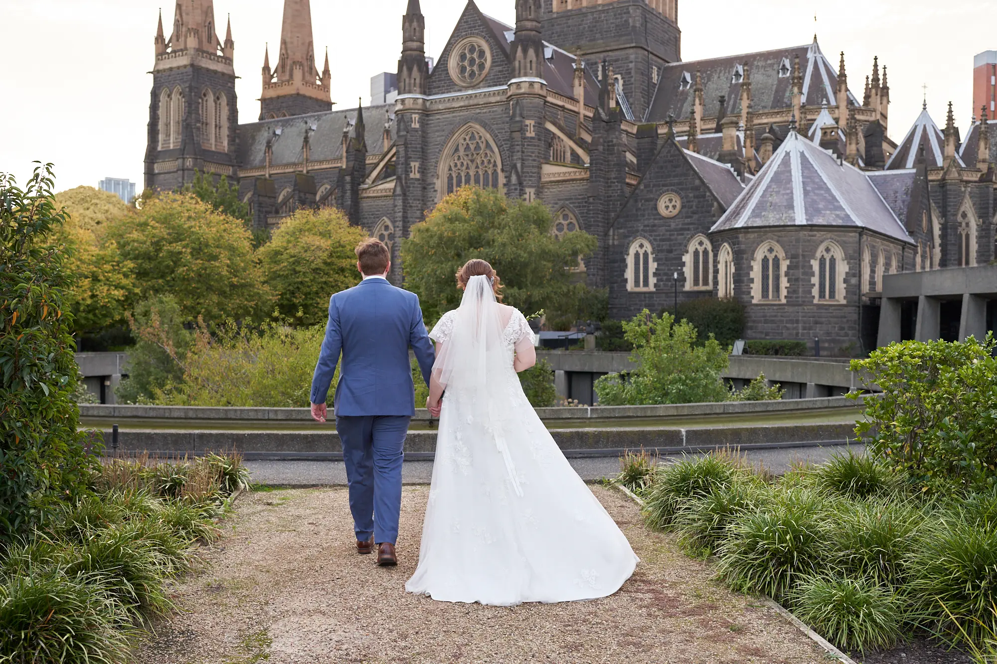 emily declan wedding bride groom garden recessional st patricks cathedral A bride and groom walk hand-in-hand away from the camera along a gravel path in a garden area with St Patrick’s Cathedral in Melbourne as the backdrop. The groom wears a light blue suit and brown shoes, and the bride wears a white V-neck A-line lace wedding dress with cap sleeves and a flowing veil. The cathedral’s dark rock-faced bluestone exterior, soaring Gothic spires, and ornate windows frame the couple, emphasizing the grandeur and historic significance of the Gothic Revival architecture. The scene conveys serene elegance and the start of their married life together.