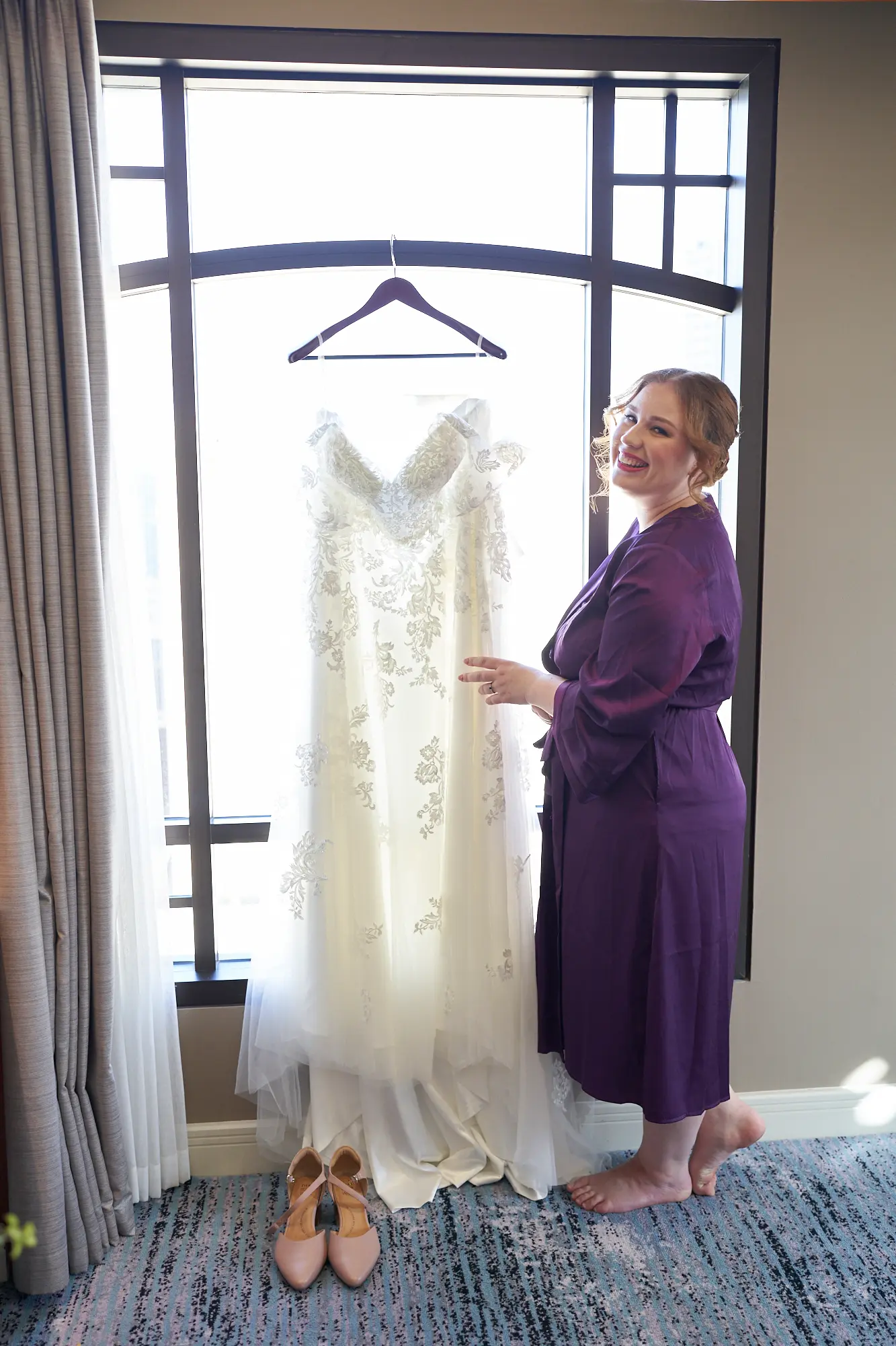 emily declan wedding bride joyful moment getting ready park hyatt melbourne The bride stands in a flowing purple robe at the Park Hyatt Melbourne, smiling warmly towards the camera as she adjusts her embroidered white wedding dress hanging nearby. Sunlight streams through a large window, illuminating the elegant hotel suite with neutral walls, patterned blue and gray carpet, and a pair of nude heels on the floor. This intimate moment captures her joyful anticipation, calm, and personal elegance, highlighting the serene atmosphere of the pre-ceremony preparations and the excitement of Emily and Declan’s wedding day.