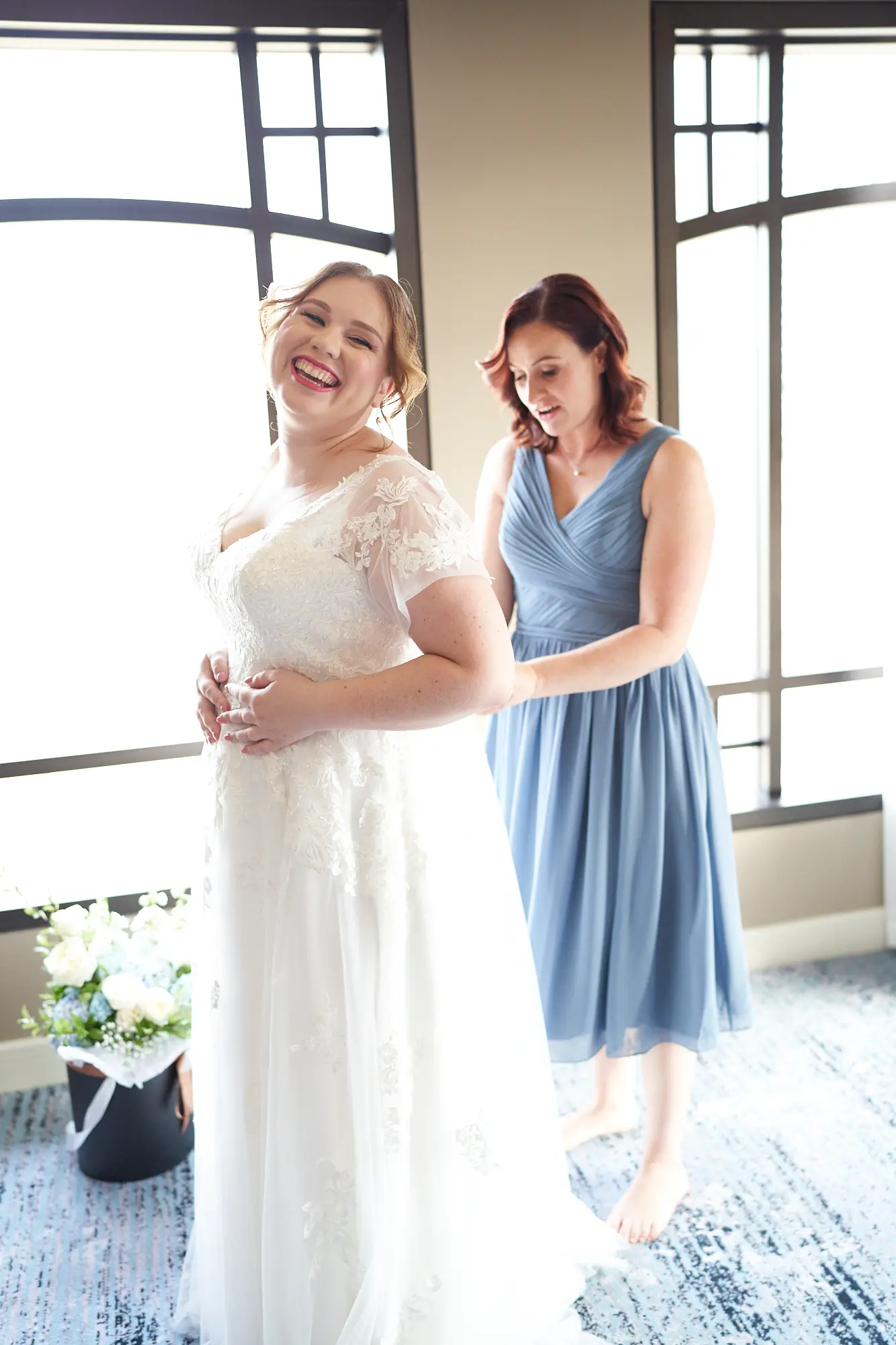 emily declan wedding bride laughing getting ready park hyatt melbourne The bride, wearing a white lace cap-sleeved wedding dress, laughs joyfully while being assisted by a bridesmaid or family member in a dusty blue dress at the Park Hyatt Melbourne. Both are barefoot on the patterned blue and white carpet, with natural light streaming through large windows, illuminating the luxurious hotel suite. A subtle floral arrangement adds elegance to the serene room. This candid moment captures warmth, anticipation, and intimate celebration, highlighting the bride’s genuine happiness and the loving support shared during Emily and Declan’s pre-ceremony preparations.