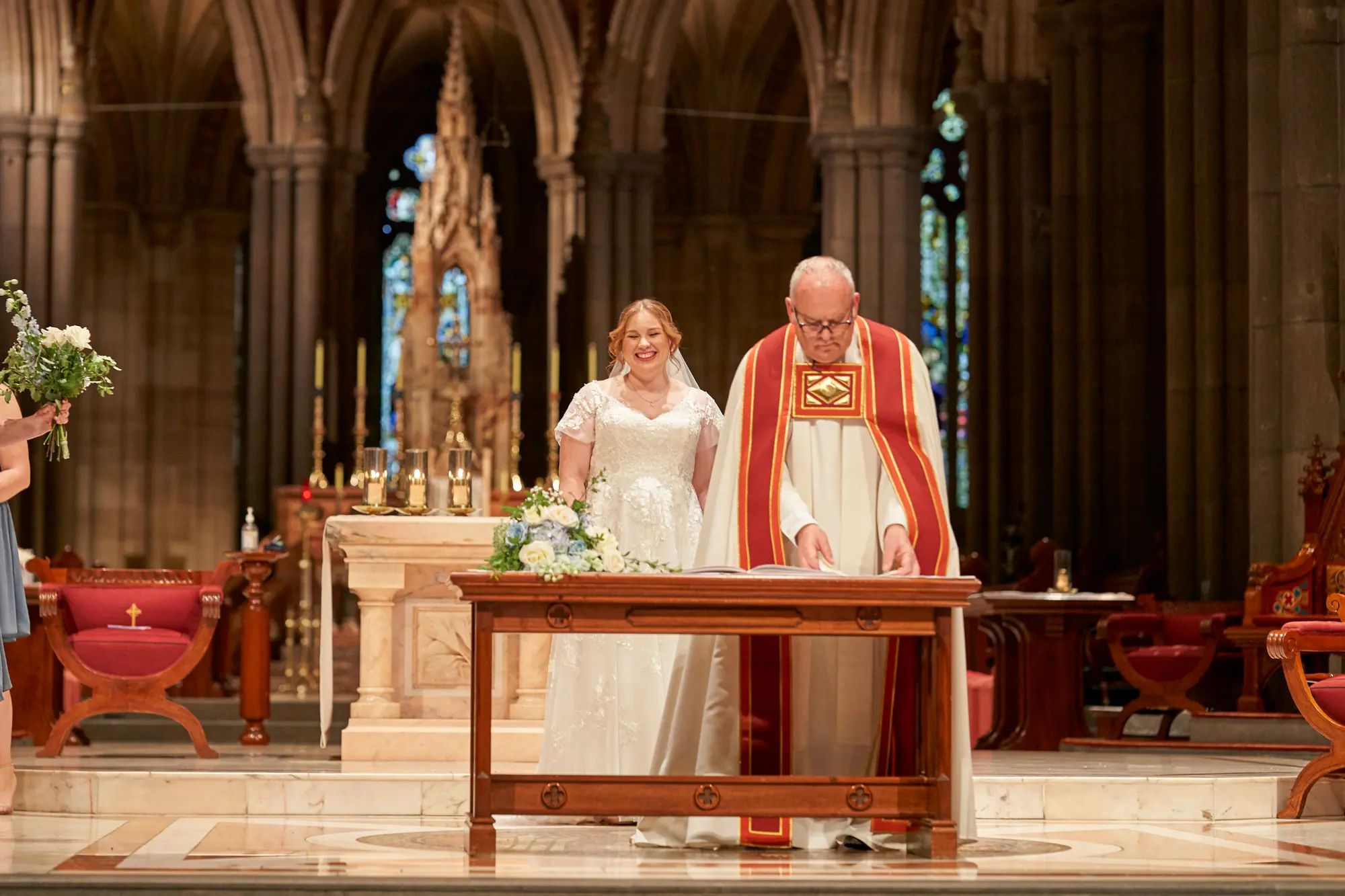 emily declan wedding bride reading ceremony st patricks cathedral melbourne The bride in a white lace gown stands at the altar of St Patrick’s Cathedral Melbourne, looking down at a red book on the lectern while smiling gently. A clergyman in white and red ceremonial vestments with an Alpha Omega Tudor Rose pattern reads from the book. A bridesmaid in a dark dress and a small child are visible nearby. The scene is framed by soaring bluestone columns, high Gothic arches, stained-glass windows, large white and green floral arrangements, candles, red chairs, and a red and white marble floor, capturing solemnity, bright reverence, and the historic grandeur of Emily and Declan’s wedding ceremony.