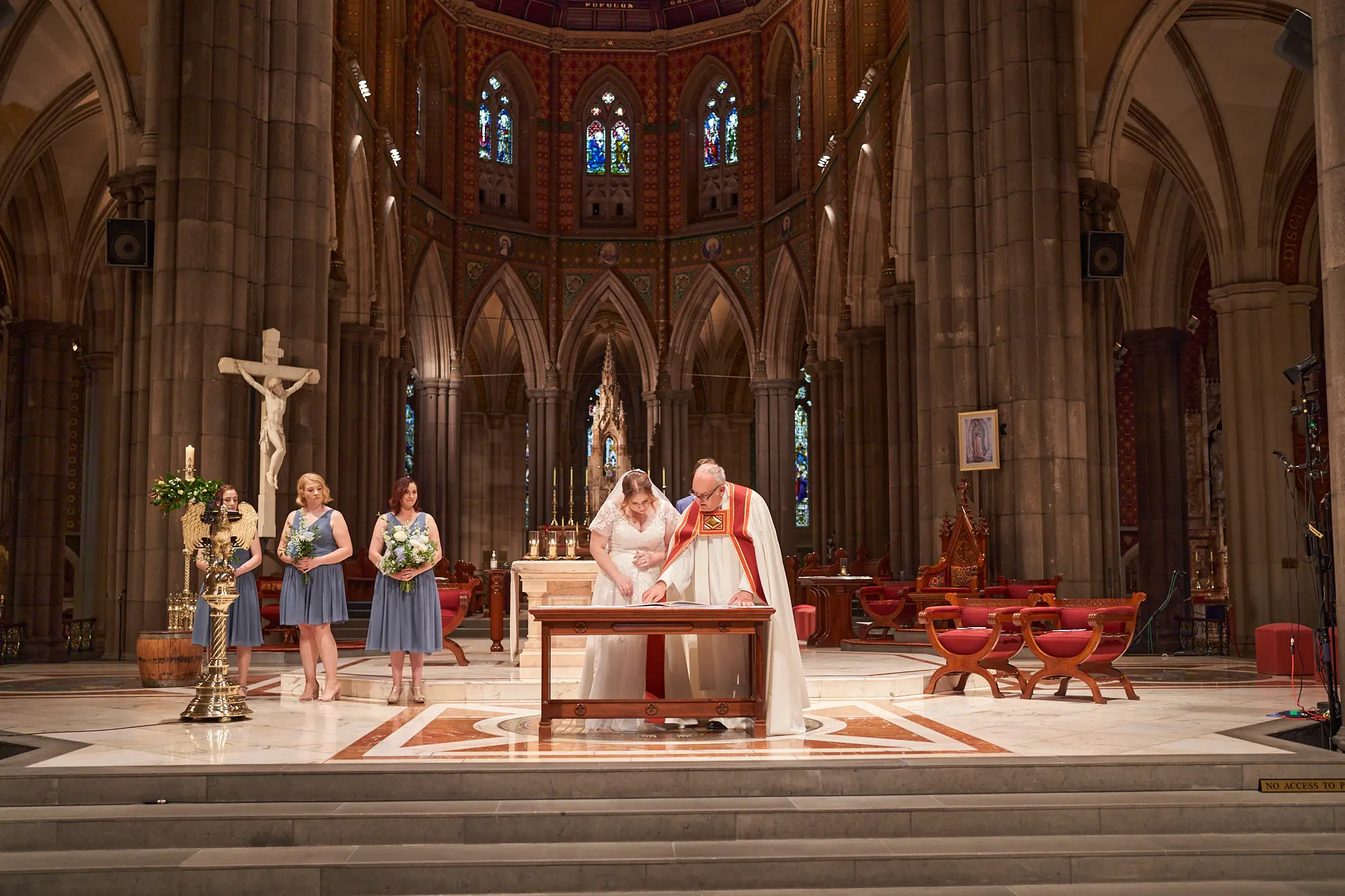 emily declan wedding bride signing certificate st patricks cathedral melbourne The bride in a white lace gown looks down as she prepares to sign the wedding certificate at St Patrick’s Cathedral Melbourne, while the groom in a blue suit and a clergyman in white and red vestments observe. Two bridesmaids in steel blue dresses stand nearby. The couple is on a raised platform with a red and white marble patterned floor, beneath soaring bluestone columns, high Gothic arches, and a mosaic-domed ceiling. Ornate stained-glass windows, a large crucifix, floral arrangements, and a gold-framed painting frame the scene, capturing solemnity, reverence, and the historic beauty of Emily and Declan’s ceremony.