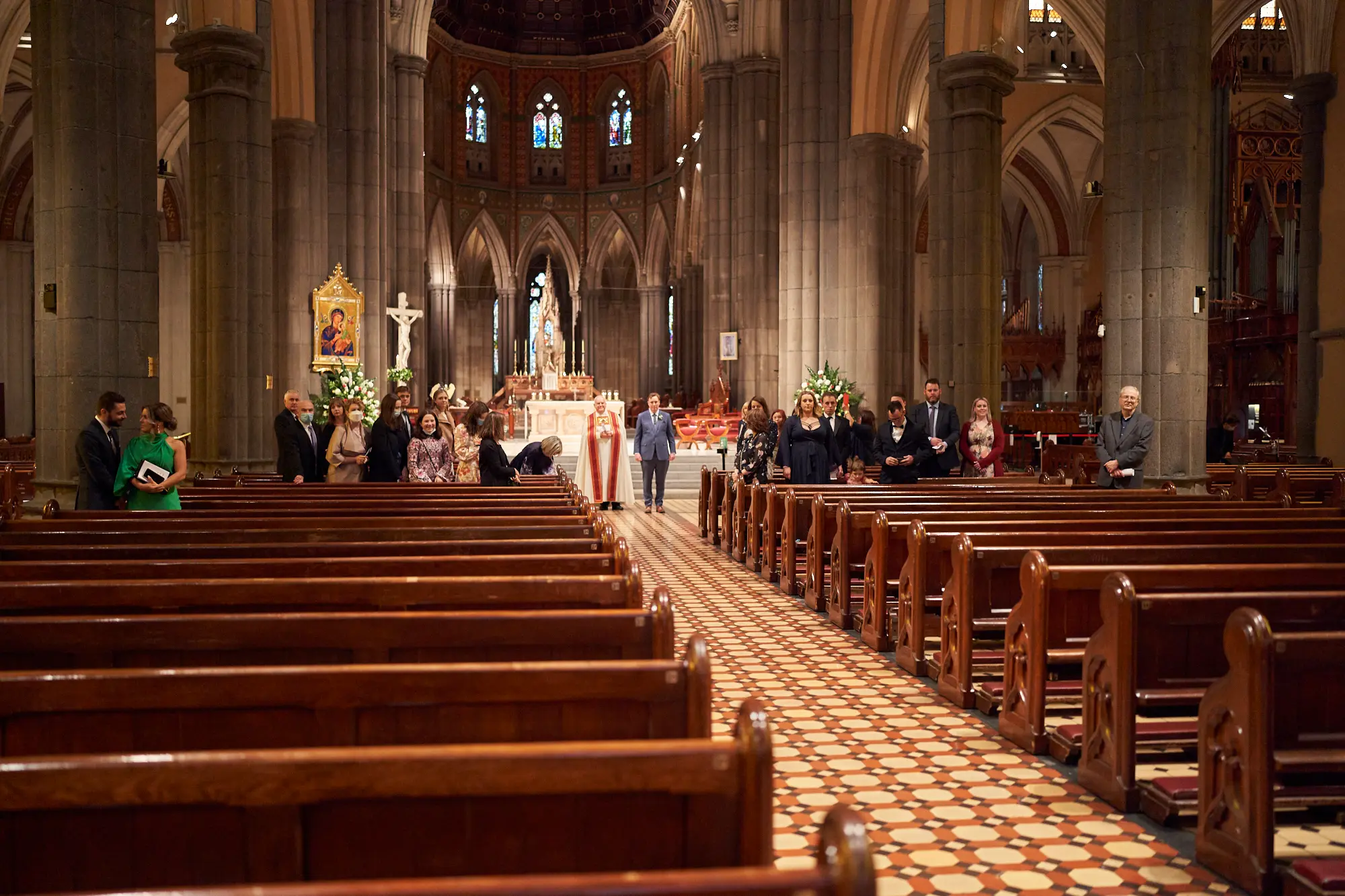 emily declan wedding ceremony altar view st patricks cathedral melbourne The wedding ceremony of Emily and Declan unfolds at St Patrick’s Cathedral Melbourne, viewed from the back of the nave down the central aisle. The groom and clergyman in red and white vestments stand at the brightly lit altar, waiting for the bride, while the wedding party and family members flank the front. Soaring bluestone pillars, high arches, and colorful stained-glass windows frame the Gothic Revival interior, with polished wooden pews and patterned tiled floors leading the eye forward. This grand, traditional moment captures solemnity, reverence, and the historic beauty of the cathedral during their sacred union.