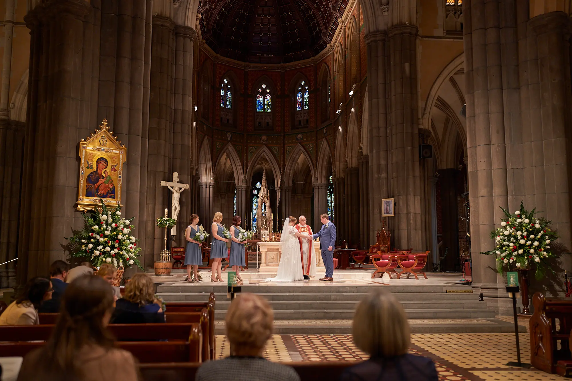 emily declan wedding ceremony bridesmaids altar st patricks cathedral melbourne The bride in a white dress and groom in a blue suit stand before a clergyman in red and white vestments at the altar of St Patrick’s Cathedral Melbourne during their wedding ceremony. Four bridesmaids in dusty blue dresses stand nearby, while the backs of seated guests are visible in the foreground. Soaring bluestone pillars, high Gothic arches, ornate mosaics, stained-glass windows, a large crucifix, and floral arrangements frame the brightly lit altar. This grand, solemn moment captures the tradition, reverence, and historic beauty of Emily and Declan’s sacred wedding ceremony.