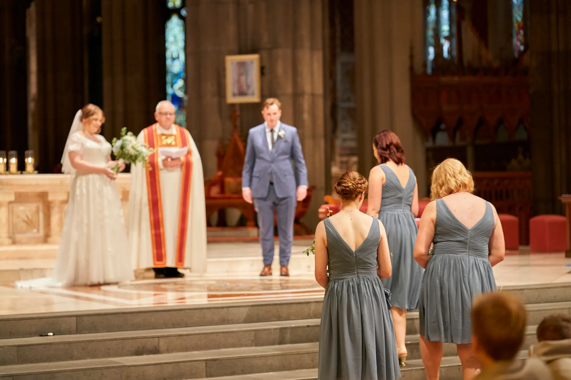 emily declan wedding ceremony wedding party altar st patricks cathedral melbourne The bride in a white lace dress, groom in a light blue suit, and a clergyman in red and white vestments stand at the altar of St Patrick’s Cathedral Melbourne during the wedding ceremony. Bridesmaids in steel blue and dusty blue dresses are seated or standing in the foreground, with guests’ heads visible in the extreme front. Soaring bluestone columns, high Gothic arches, colorful stained-glass windows, and white and green floral arrangements frame the brightly lit altar. This grand, traditional moment captures solemnity, reverence, and the historic beauty of Emily and Declan’s sacred wedding ceremony.