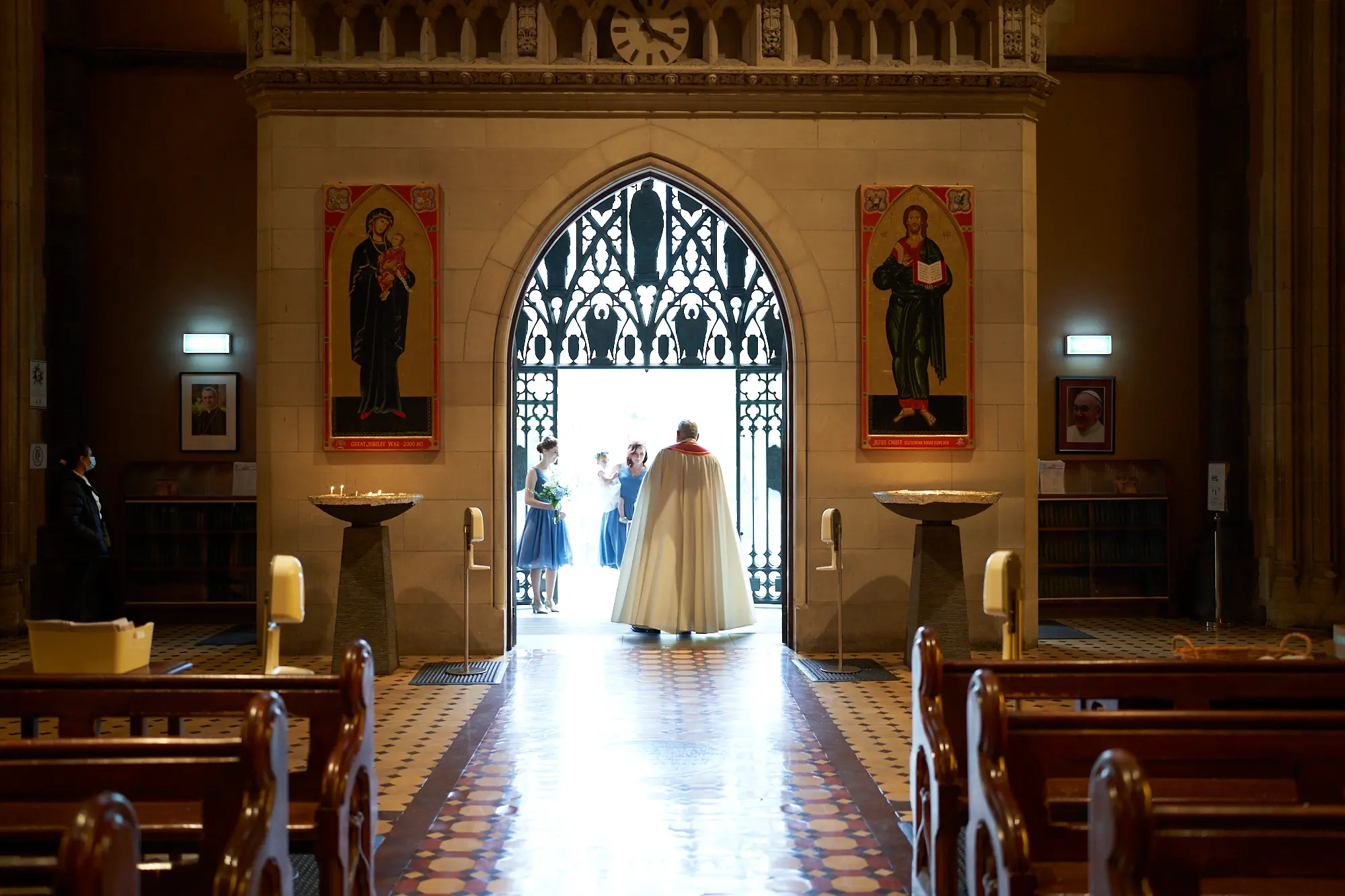 emily declan wedding clergyman entrance st patricks cathedral melbourne A clergyman in a white and red ceremonial cape and mitre walks through the main doors of St Patrick’s Cathedral Melbourne, leading the wedding procession. Behind him, bridesmaids in light blue dresses and other guests await their turn to enter. The grand stone archway, intricate iron gates, and bluestone pillars frame the scene, while patterned tiled floors and dark wooden pews emphasize the cathedral’s Gothic Revival architecture. Natural light streams through the doorway, contrasting with the dim interior and creating a solemn, anticipatory atmosphere, capturing the grandeur and reverence of Emily and Declan’s wedding ceremony entrance.