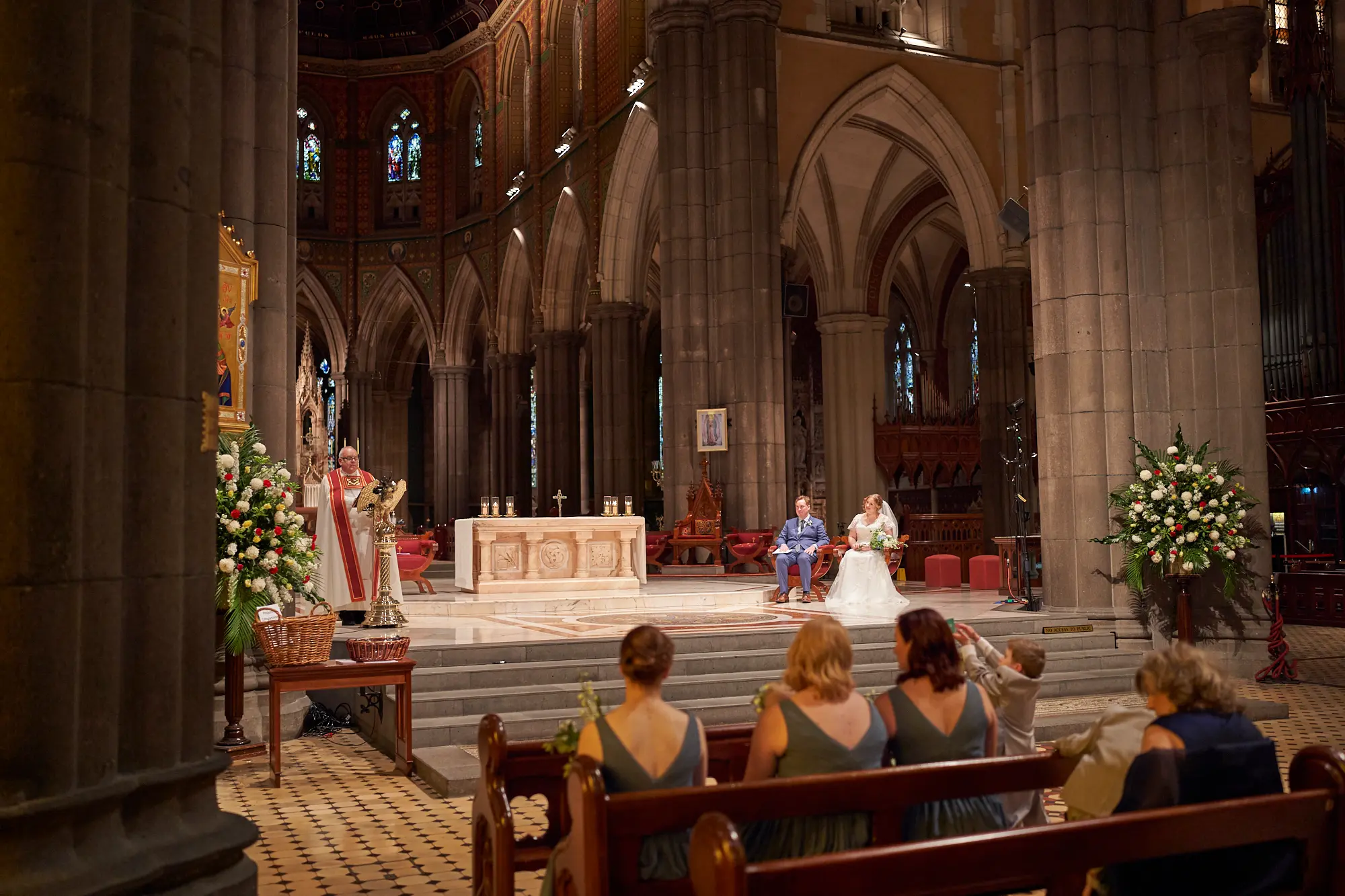 emily declan wedding couple seated ceremony st patricks cathedral melbourne The bride and groom are seated at the altar of St Patrick’s Cathedral Melbourne during their wedding ceremony, accompanied by the maid of honor and best man. A clergyman in white and red vestments stands at a brass eagle lectern addressing the congregation. Guests are seated in dark wooden pews in the foreground, some visible from behind. Soaring bluestone columns, high Gothic arches, ornate stained-glass windows, mosaics, a marble altar, and a white alabaster canopy frame the brightly lit sanctuary. This grand, traditional moment captures solemnity, reverence, and the historic beauty of Emily and Declan’s sacred wedding ceremony.
