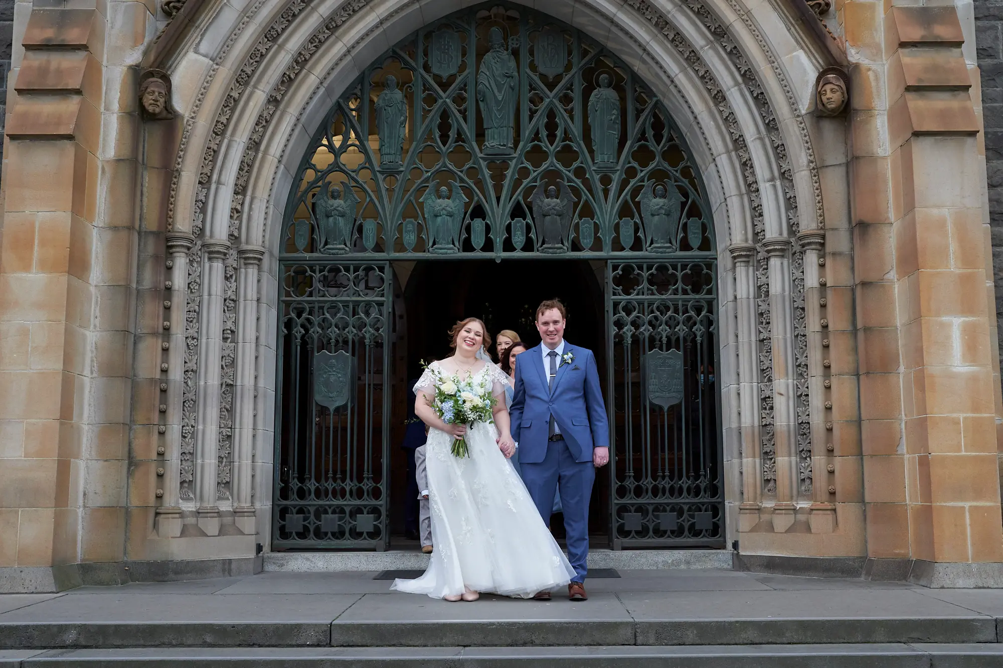 emily declan wedding exit st patricks cathedral melbourne The bride in a white lace wedding dress holding a white and blue bouquet and the groom in a light blue suit and brown shoes walk hand-in-hand down the steps of St Patrick’s Cathedral Melbourne, beaming with joy after their ceremony. Behind them, members of the wedding party follow. The grand Gothic Revival entrance with intricate stone carvings, decorative wrought-iron gates, and a large open archway frames the scene, creating a bright, celebratory, and elegant atmosphere as the couple exits into the sunlight.