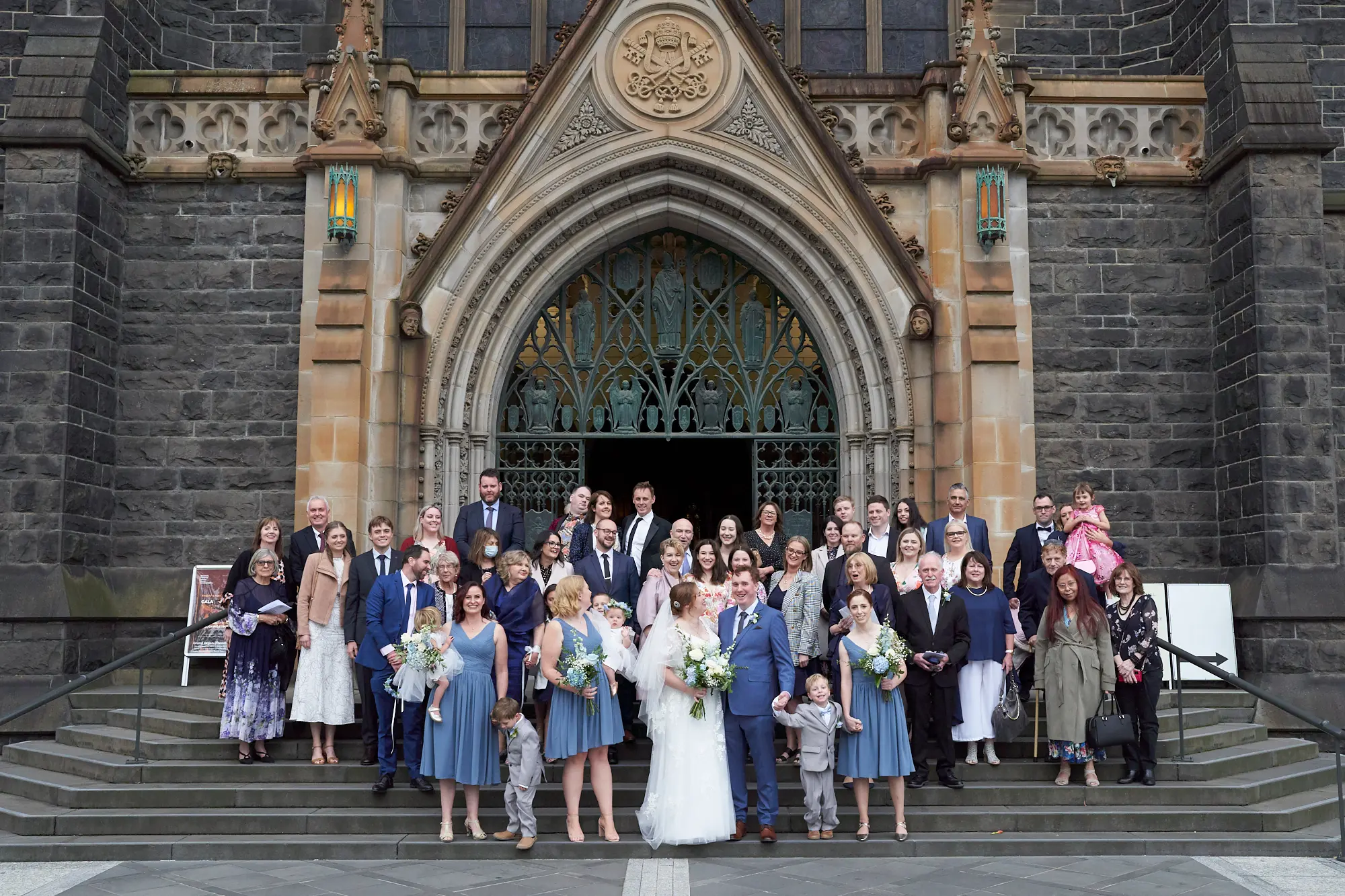 emily declan wedding family group st patricks cathedral melbourne A large wedding group of approximately 40 people, including the bride in a white lace dress with cap sleeves holding a white and blue bouquet and the groom in a light blue suit, is posed on the wide stone steps of St Patrick’s Cathedral Melbourne. Bridesmaids in dusty blue dresses and groomsmen in blue suits flank the couple, while family members and guests, including young children in formal attire, are arranged on the steps above and beside them. The historic bluestone Gothic Revival facade, featuring an ornate stone archway, intricate iron grille, sculptures, and the Papal Coat of Arms, provides a grand and elegant backdrop, creating a joyous and celebratory group portrait.