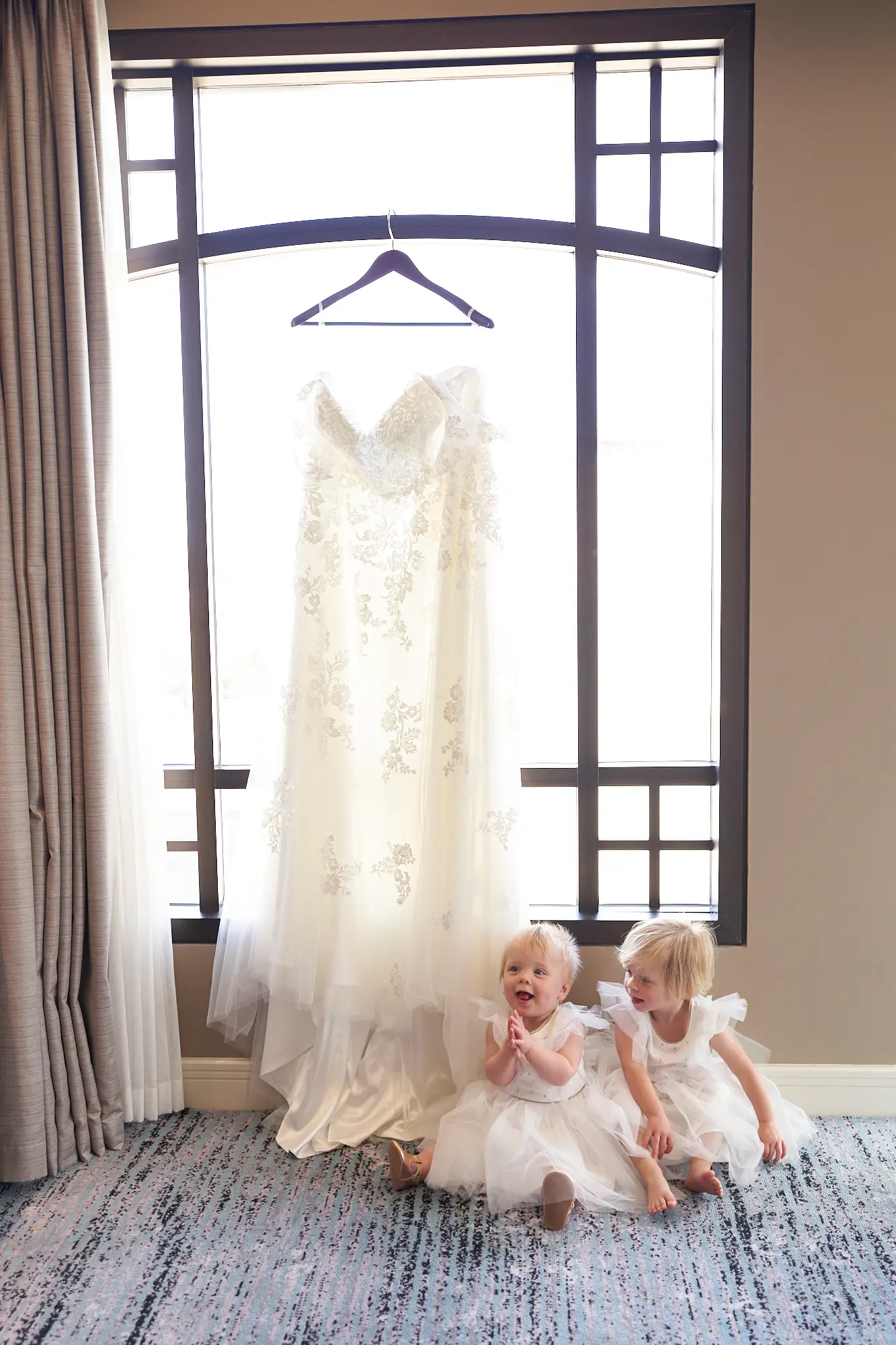 emily declan wedding flower girls bride getting ready park hyatt melbourne Two young flower girls in matching white tulle dresses sit on the carpeted floor of a Park Hyatt Melbourne suite, sharing a joyful moment while the bride’s lace and beaded wedding gown hangs above in the window. Soft natural light illuminates the elegant room, highlighting neutral tones, patterned carpet, and luxurious details. Their candid expressions and playful gestures contrast with the poised bridal gown, capturing the innocence, excitement, and quiet elegance of the pre-wedding preparation, reflecting the intimate and heartfelt atmosphere of Emily and Declan’s wedding day.