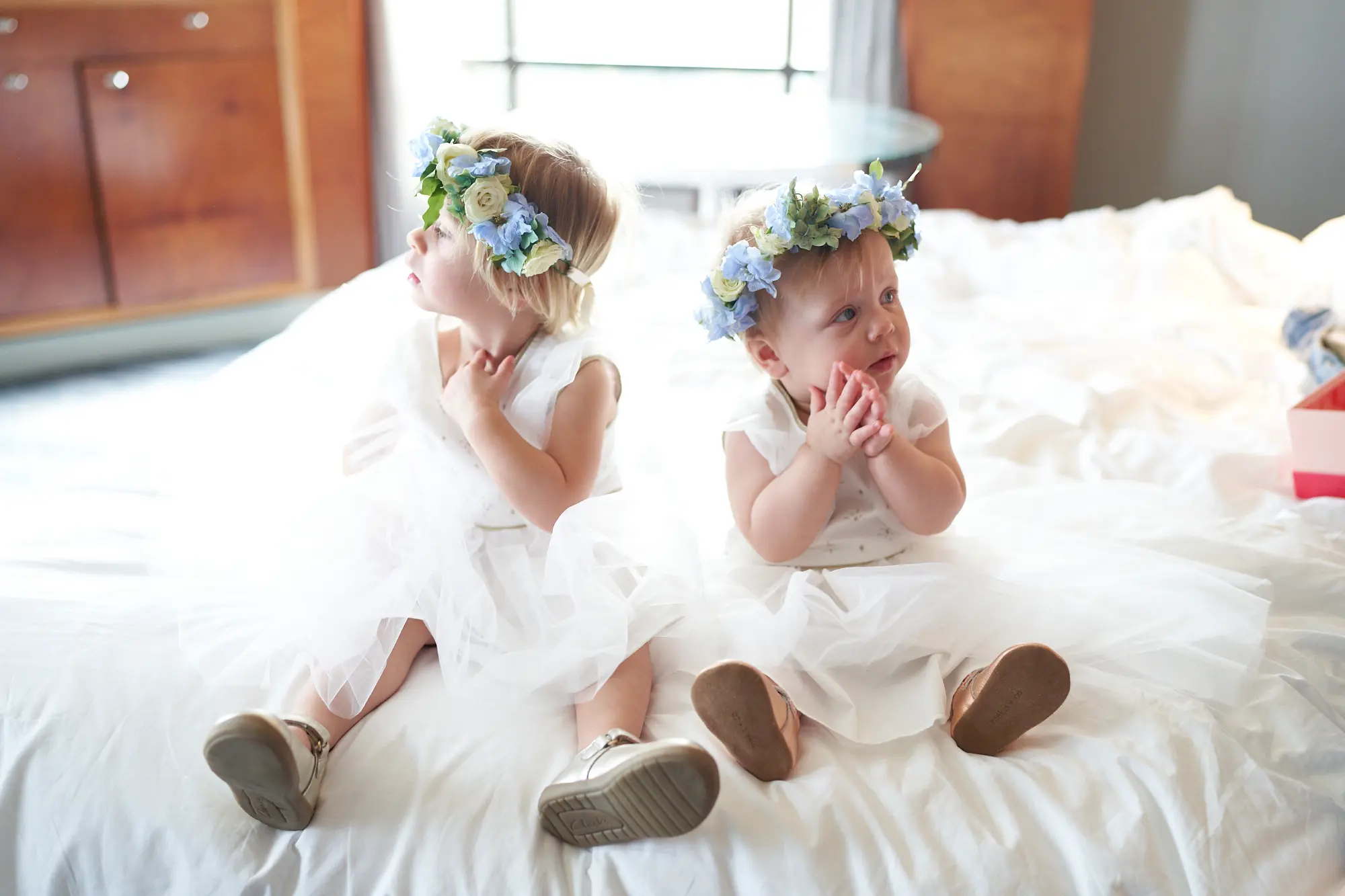 emily declan wedding flower girls preparation park hyatt melbourne Two young flower girls in white dresses with delicate blue and green floral crowns sit on a plush white bed at the Park Hyatt Melbourne, captured in a candid moment of innocence and anticipation. One girl clasps her hands at her chest while the toddler beside her looks upward thoughtfully. Natural light fills the bright hotel suite, highlighting the white bedding and soft textures, while dark wood furniture adds subtle elegance. This heartwarming scene reflects the gentle excitement and serene atmosphere of Emily and Declan’s pre-ceremony wedding preparations.