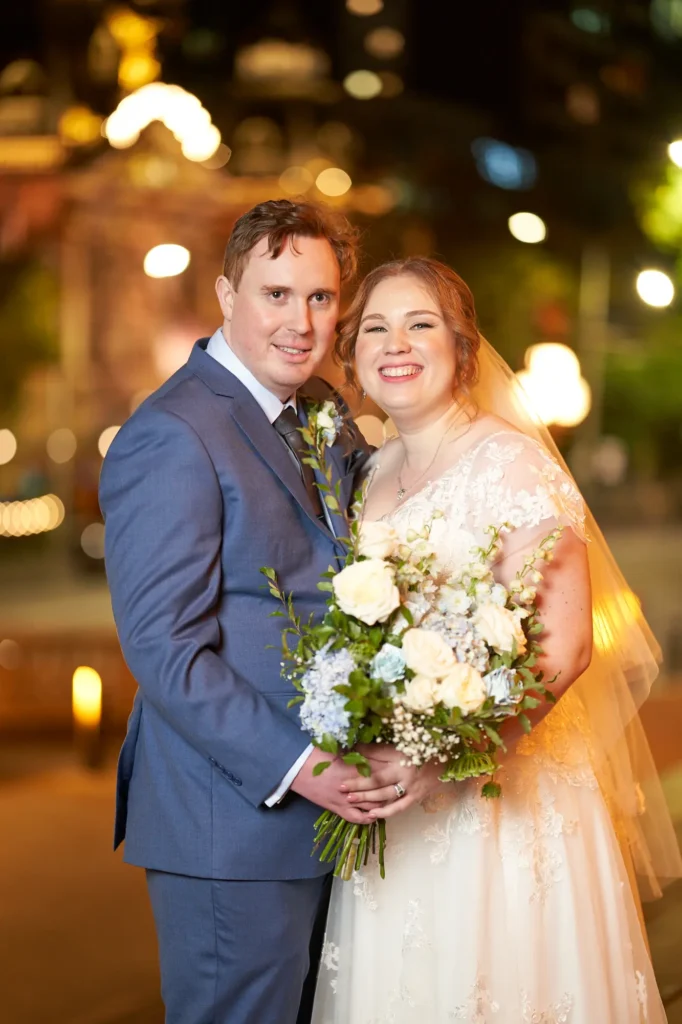 emily declan wedding melbourne city night portrait A joyful nighttime wedding portrait of a bride and groom standing closely together with warm, softly blurred city lights behind them, including the Princess Theatre Melbourne. The groom wears a light blue suit, and the bride wears a white V-neck lace gown with a flowing veil while holding a white and blue bouquet. They smile brightly at the camera, creating a vibrant, romantic, and cinematic moment.