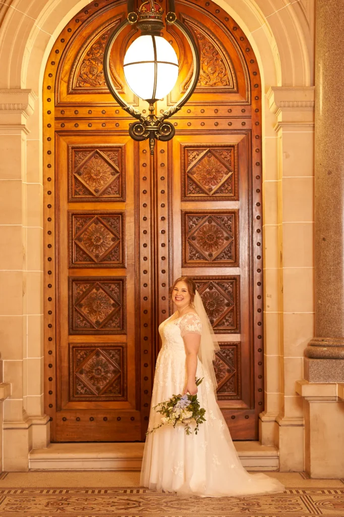 emily declan wedding parliament house melbourne bride portrait A formal bridal portrait taken at the grand entrance of Parliament House Melbourne, featuring the bride smiling warmly in a white V-neck lace gown with cap sleeves and a mid-length veil. She holds a bouquet of white and blue flowers while standing before ornate carved wooden doors, polished red granite columns, and a glowing bronze light fixture, creating an elegant and historic atmosphere.