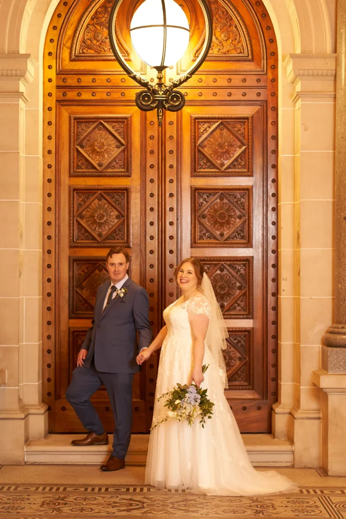 emily declan wedding parliament house melbourne couple laughing portrait A joyful portrait of the bride and groom at the grand entrance of Parliament House Melbourne. The couple stand close together, laughing warmly at the camera while holding a bouquet of white and blue flowers. The groom lifts his left leg slightly on the step, adding dynamic movement, while the bride stands in her V-neck lace gown with flowing veil. Behind them are richly carved wooden doors, polished red granite columns, and a glowing bronze light fixture, creating an elegant and historic backdrop.
