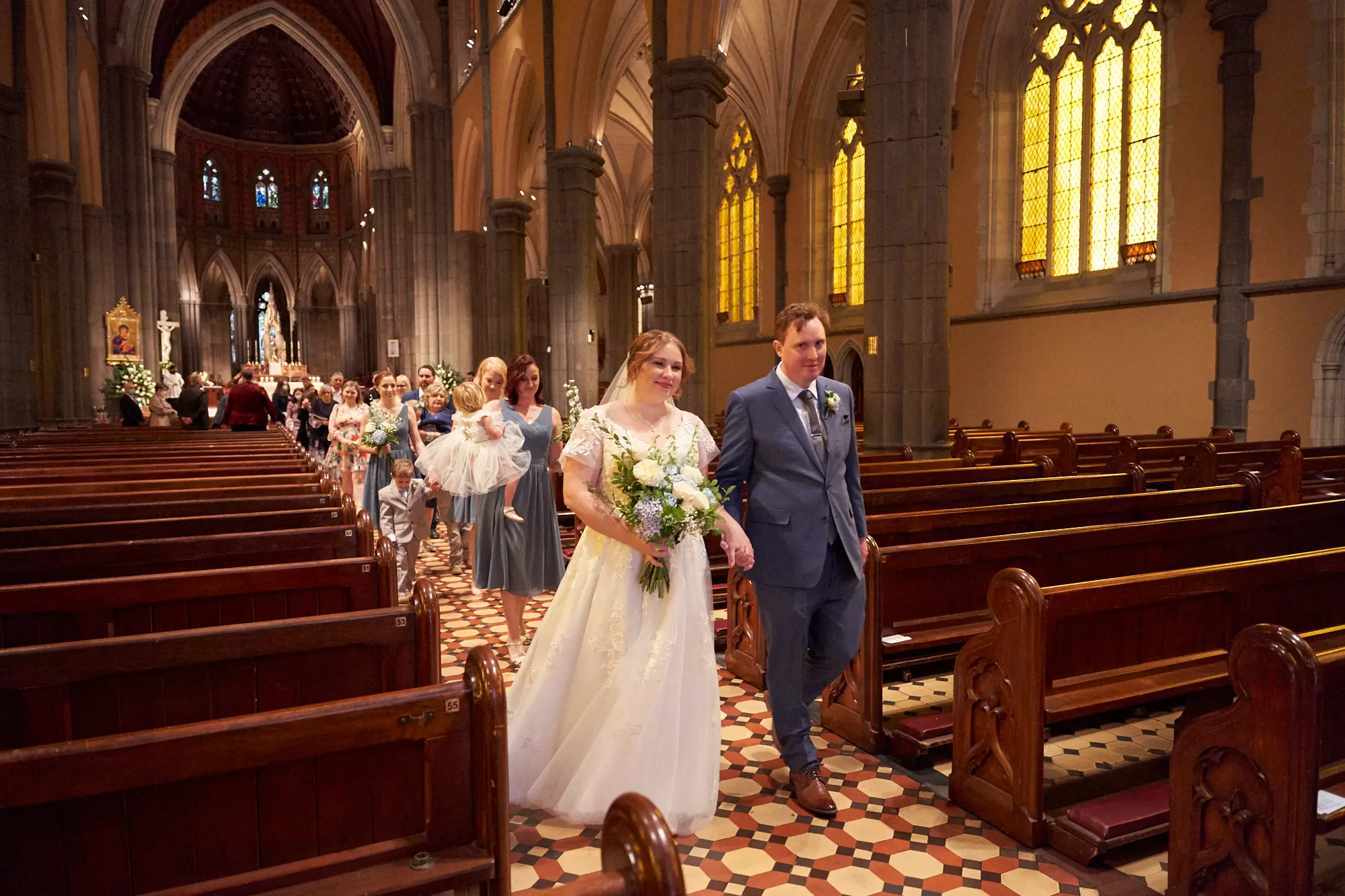 emily declan wedding recessional aisle procession st patricks cathedral melbourne The bride in a white lace wedding dress holding a large bouquet and the groom in a blue suit walk hand-in-hand down the central aisle of St Patrick’s Cathedral Melbourne after their ceremony. Bridesmaids in dusty blue dresses, a ring bearer, and a flower girl follow behind. Guests watch from dark wooden pews as the Gothic Revival interior with soaring bluestone columns, high arches, red-and-white patterned aisle tiles, a brightly lit altar with crucifix, and ornate stained-glass windows provides a grand, joyful, and reverent backdrop.