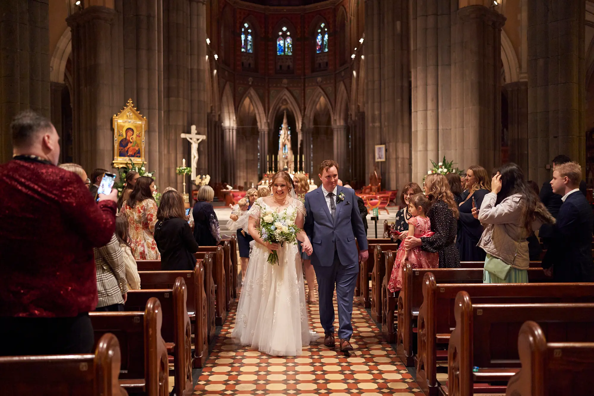 emily declan wedding recessional aisle st patricks cathedral melbourne The bride in a white lace wedding dress and groom in a blue suit walk hand-in-hand down the central aisle of St Patrick’s Cathedral Melbourne after their wedding ceremony. Guests line the pews, some smiling and taking photos, while a small child in pink watches. The Gothic Revival interior features soaring bluestone columns, high arches, red and white patterned aisle tiles, a brightly lit altar with a large crucifix, ornate stained-glass windows, and dark wooden pews. The scene conveys joy, celebration, and timeless elegance as the newlyweds recess together.
