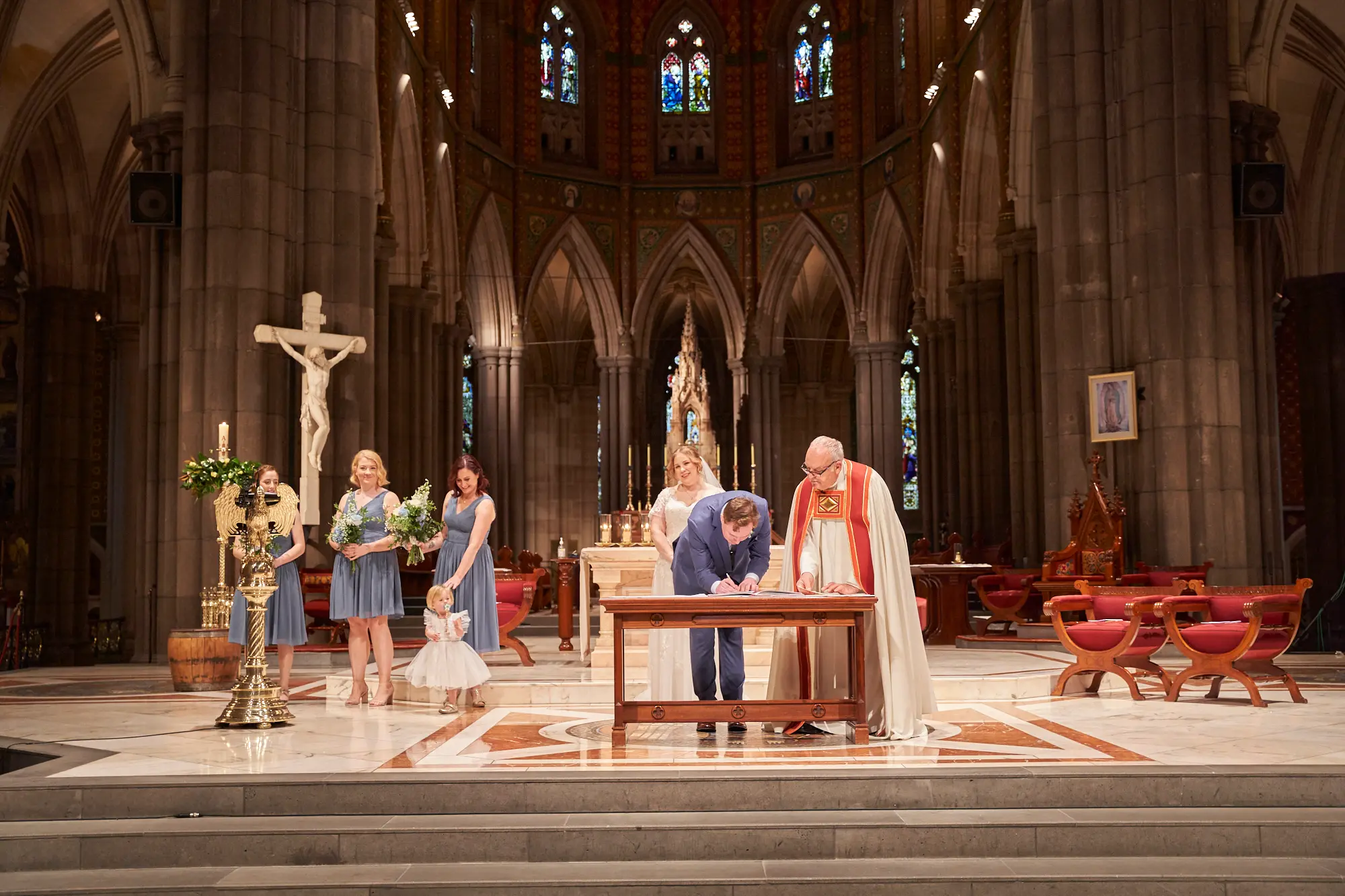 emily declan wedding signing ceremony st patricks cathedral melbourne The groom in a blue suit bends over to sign one of the required marriage documents at a small wooden table on the altar of St Patrick’s Cathedral Melbourne, while the bride in her white lace gown stands nearby holding her bouquet, waiting her turn. A clergyman in red and white vestments observes, and several bridesmaids in dusty blue dresses along with two young flower girls in white stand attentively to the side. Soaring bluestone columns, high Gothic arches, stained-glass windows, a large crucifix, religious paintings, floral arrangements, and a red and white marble floor frame this solemn, historic, and reverent moment of Emily and Declan’s wedding ceremony.