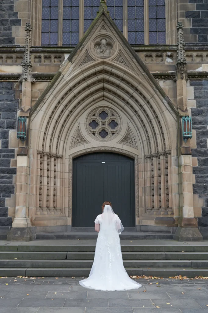 emily declan wedding st patricks cathedral bride entrance This image captures a bride posing for a solo portrait at the entrance of St Patrick's Cathedral in Melbourne, viewed from behind. She wears an elegant white lace wedding gown with cap sleeves and a flowing train, along with a long, sheer veil draping across the stone steps. The dark bluestone Gothic Revival cathedral with ornate arches, carved details, and tall tracery windows creates a grand, historic backdrop. Fallen autumn leaves on the paved courtyard add natural texture, enhancing the serene, contemplative, and timeless atmosphere of this pre-ceremony wedding moment.