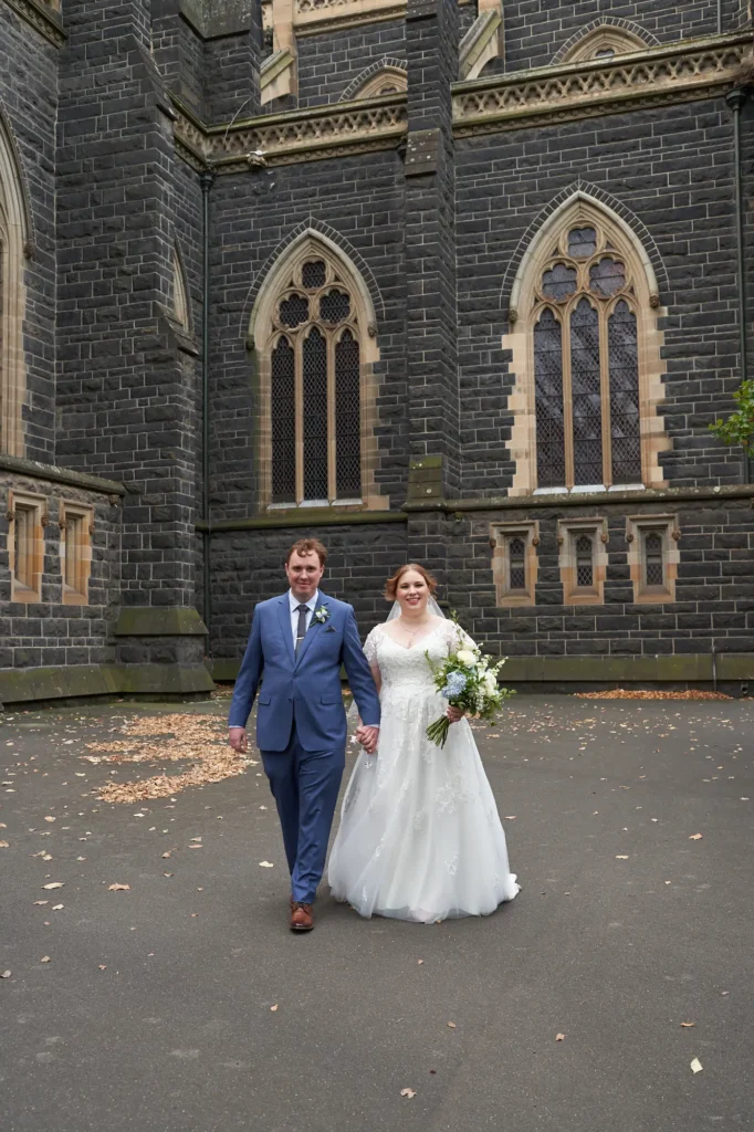 emily declan wedding st patricks cathedral courtyard entrance This image captures a bride and groom walking hand-in-hand towards the camera in a paved courtyard, with St Patrick's Cathedral in Melbourne as a dramatic historic backdrop. The bride wears a white V-neck A-line lace dress with cap sleeves and holds a large bouquet of white and blue flowers. The groom wears a light blue suit with a patterned tie. The Gothic Revival bluestone cathedral, ornate pointed-arch windows, and sandstone trim provide a majestic contrast to the joyful couple. Fallen autumn leaves and low green foliage add natural texture, creating a serene, elegant, and timeless wedding moment.